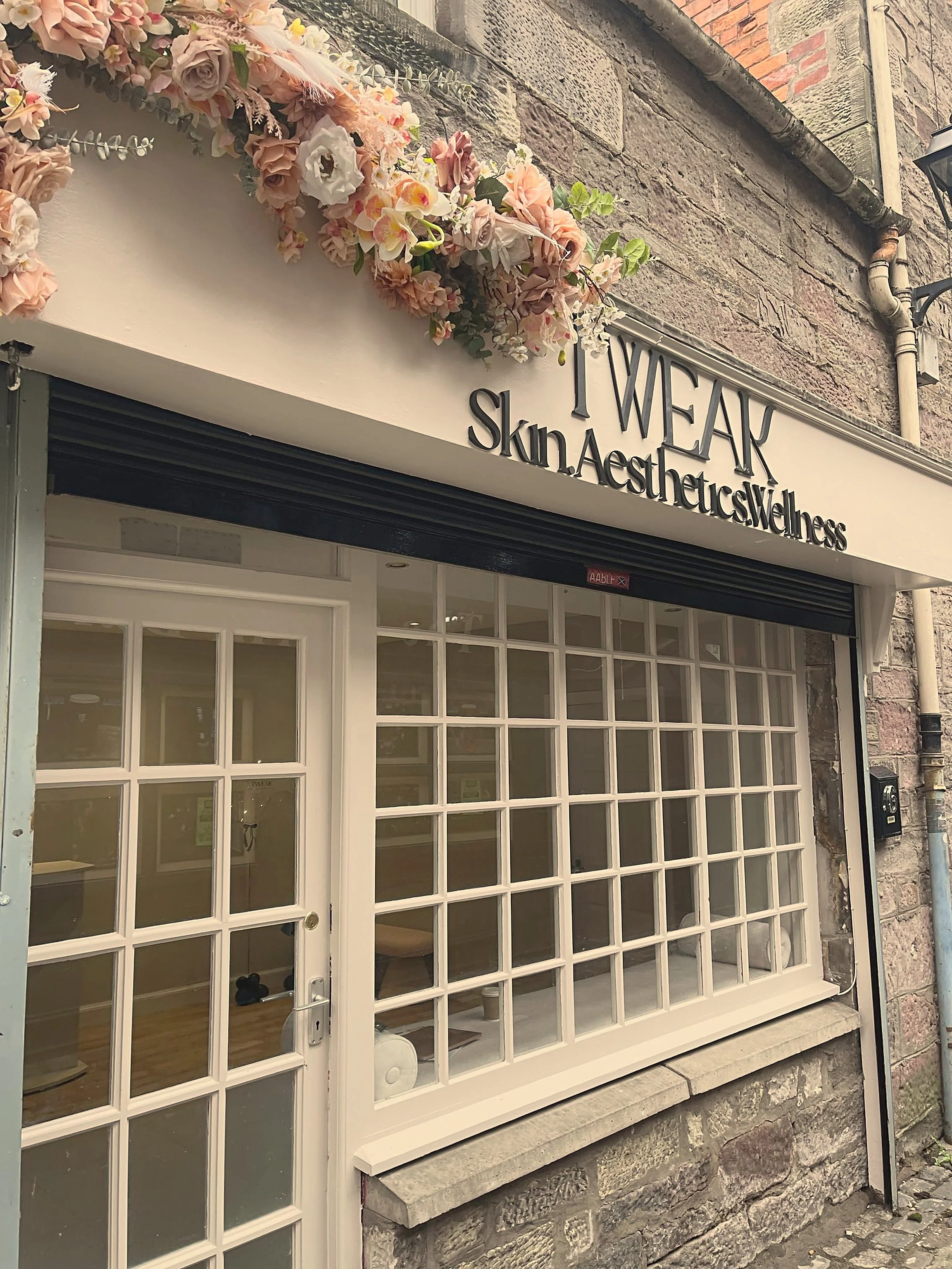 A storefront with a decorative wooden sign that reads "TWEAK" and "skin & aesthetic wellbeing." There is a large white-framed window with multiple panes and a door, and above it is a floral arrangement of pink and white flowers and greenery. The building has a brick and stone exterior with a pipe and a black outdoor lamp.