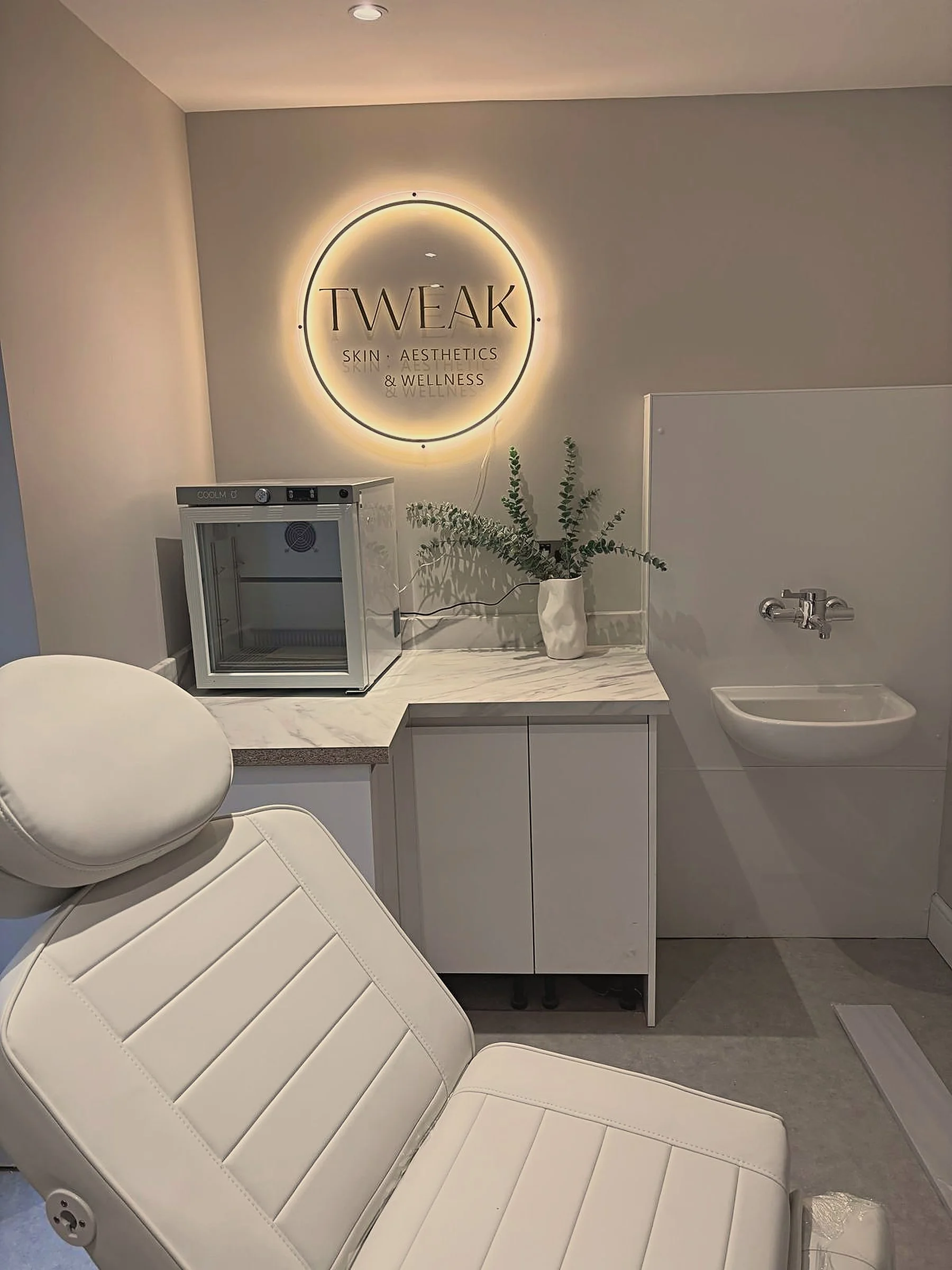 A medical or spa treatment room with a white reclining chair, a small white cabinet with a mini fridge, a potted plant, and a wall sign that reads "TWEAK Skin, Aesthetics & Wellness" illuminated by backlighting.