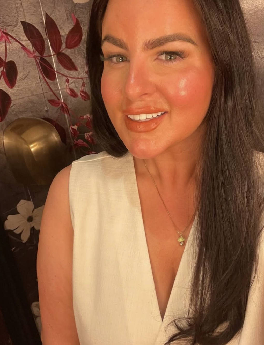 Close-up of a woman with long brown hair, wearing a cream sleeveless top, a gold necklace with a flower pendant, smiling in front of a floral background with red leaves and white flowers.
