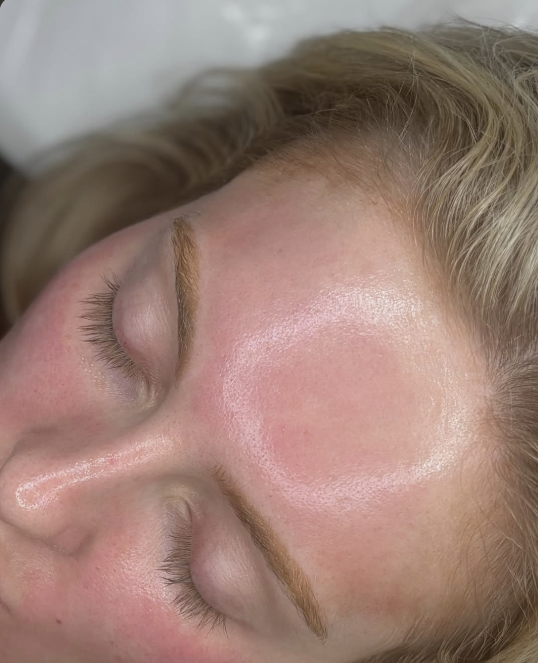 Close-up of a woman's forehead, eyebrows, and part of her eyes with natural eyelashes, showing smooth skin and a shiny, dewy complexion.
