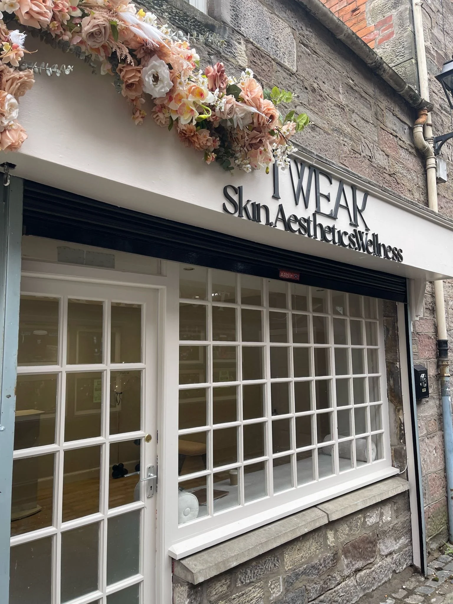 Front view of a shop with a white door and large grid-patterned windows, decorated with pink and white floral arrangements above the door, and a stone wall to the side with a black mailbox and a street lamp.