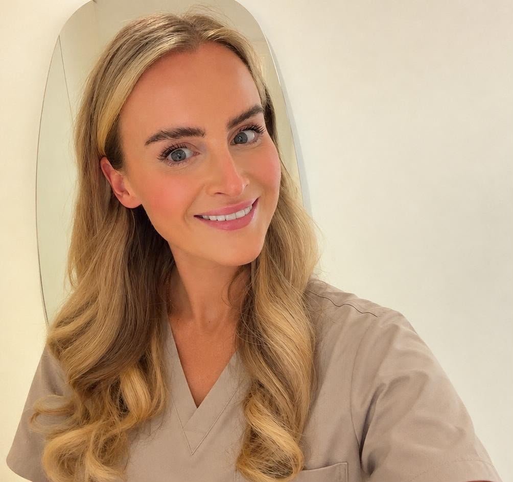 A woman with long, wavy blonde hair taking a selfie in front of an oval mirror, smiling, wearing a beige top, against a plain light-colored wall.