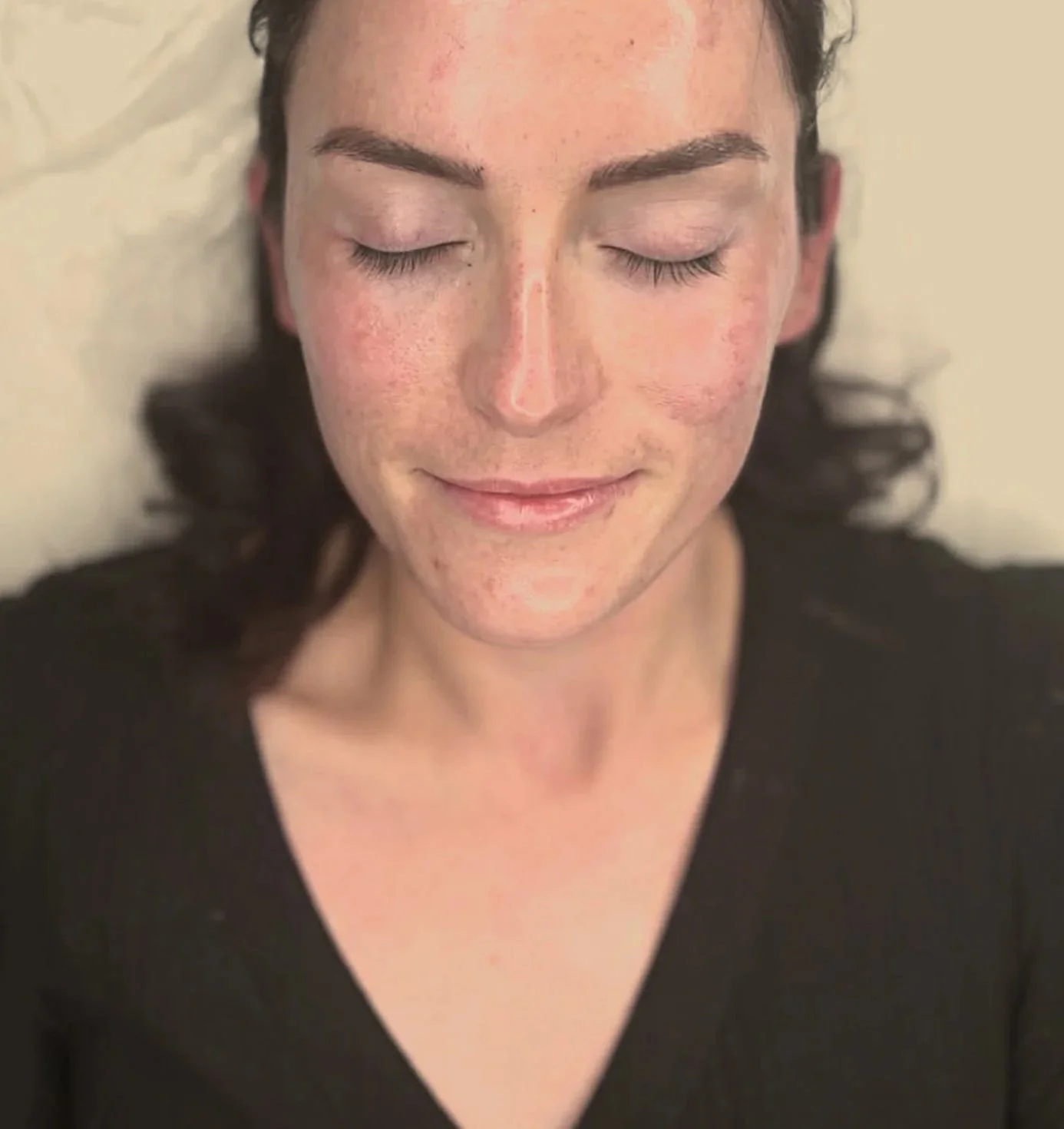 A woman with dark curly hair and light skin, eyes closed, smiling softly, wearing a black V-neck top, resting on a light-colored pillow or surface.