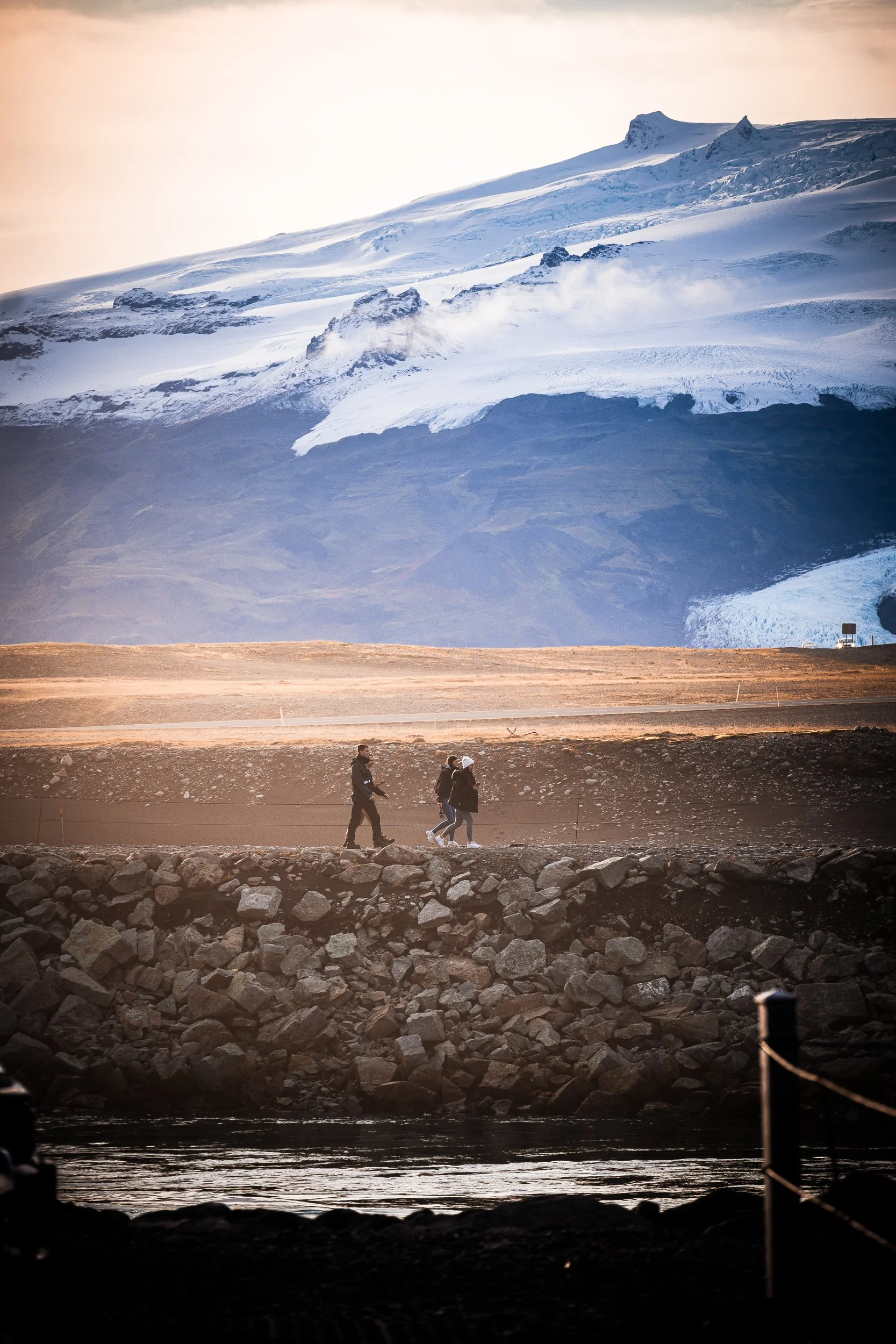 Hikers walking beside a road beneath a snow-covered glacier and volcanic mountain