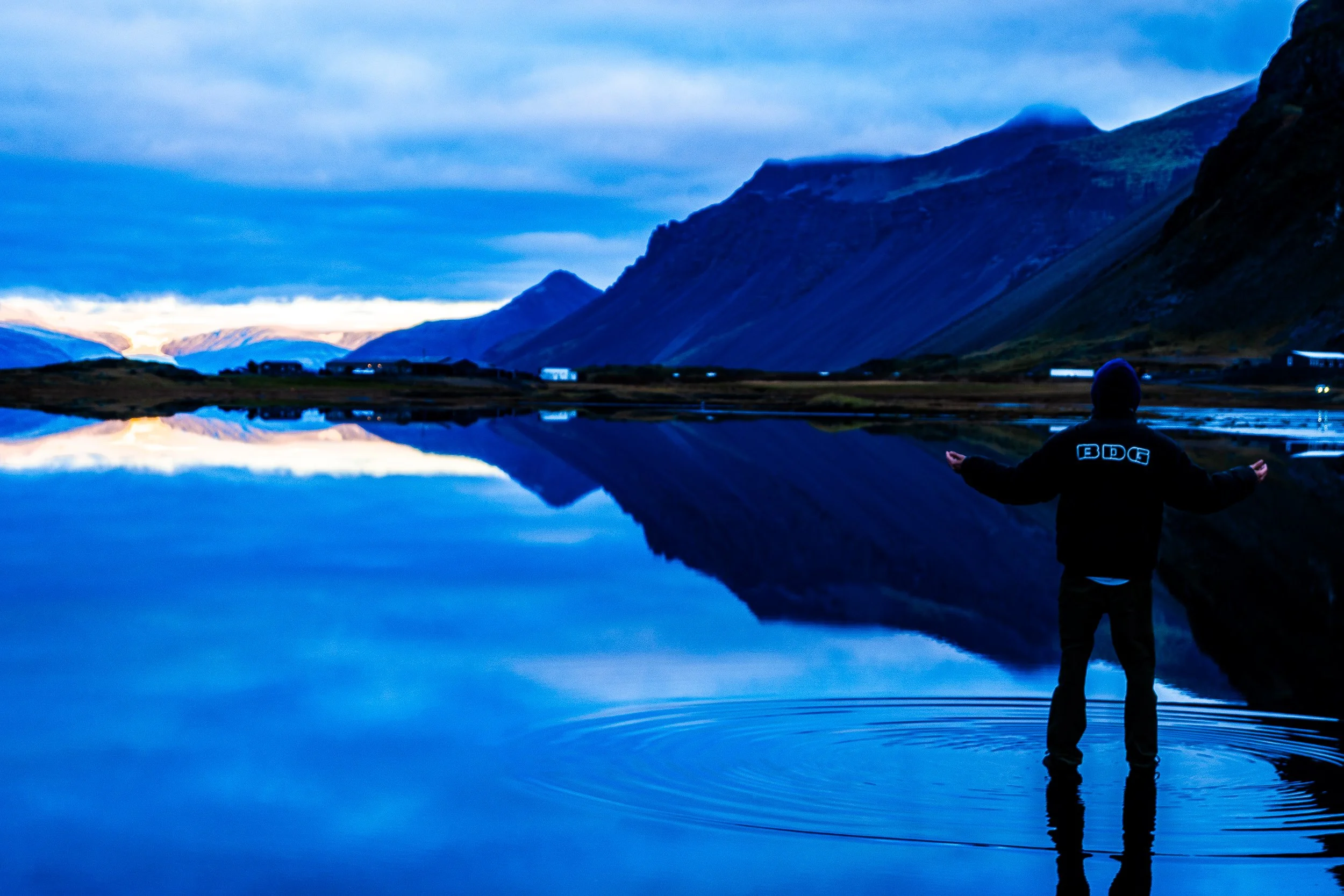 Person standing in a calm mountain lake at dusk, surrounded by dramatic peaks reflected in the water