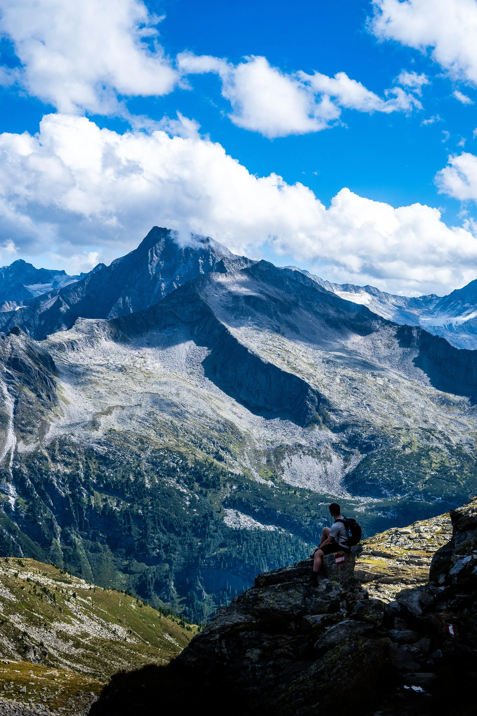 Hiker sitting on rocky mountain slope overlooking rugged, snow-dusted peaks beneath a bright blue sky