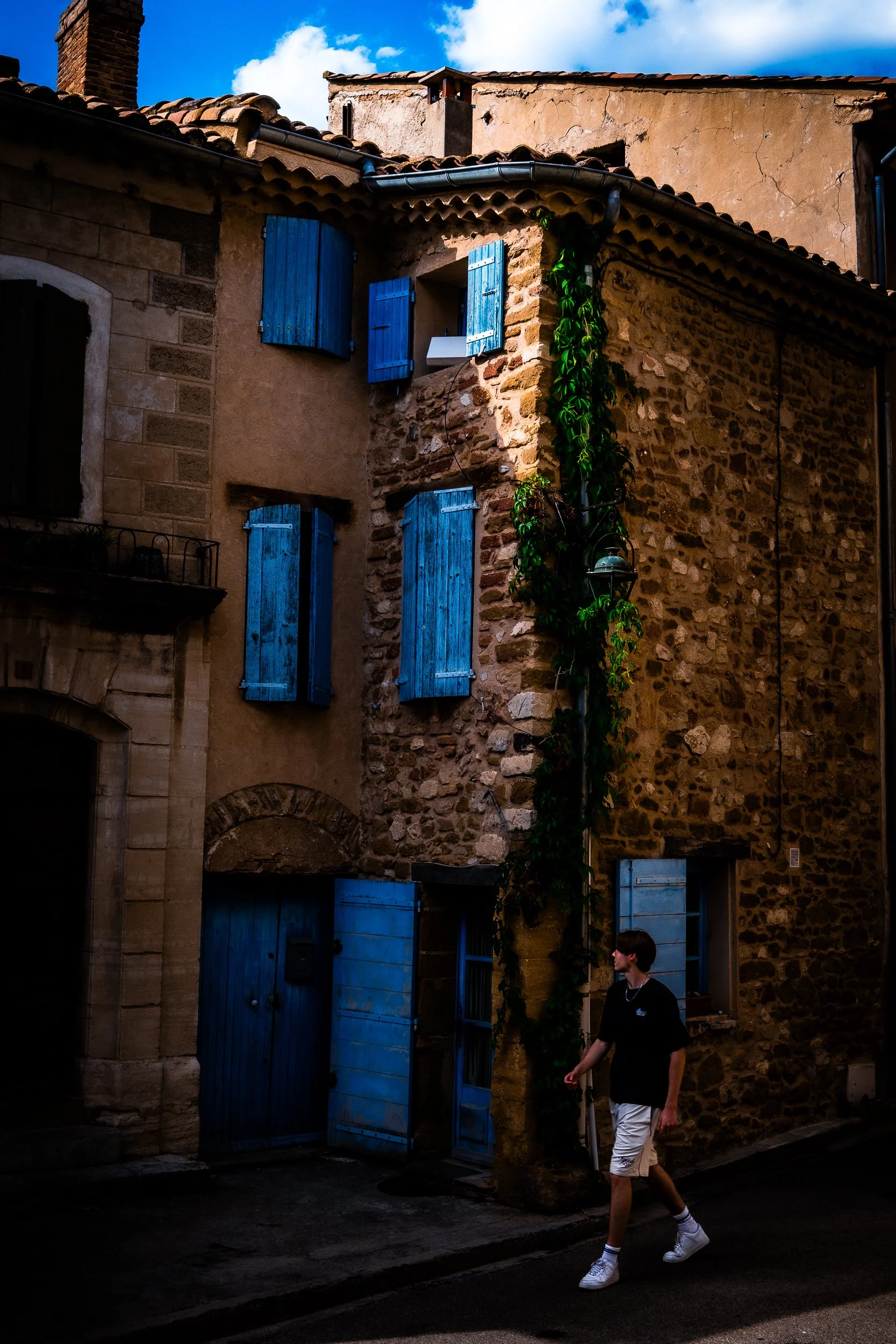 Person walking past a historic stone-and-stucco building with blue shutters, tiled roofs, and climbing vines.