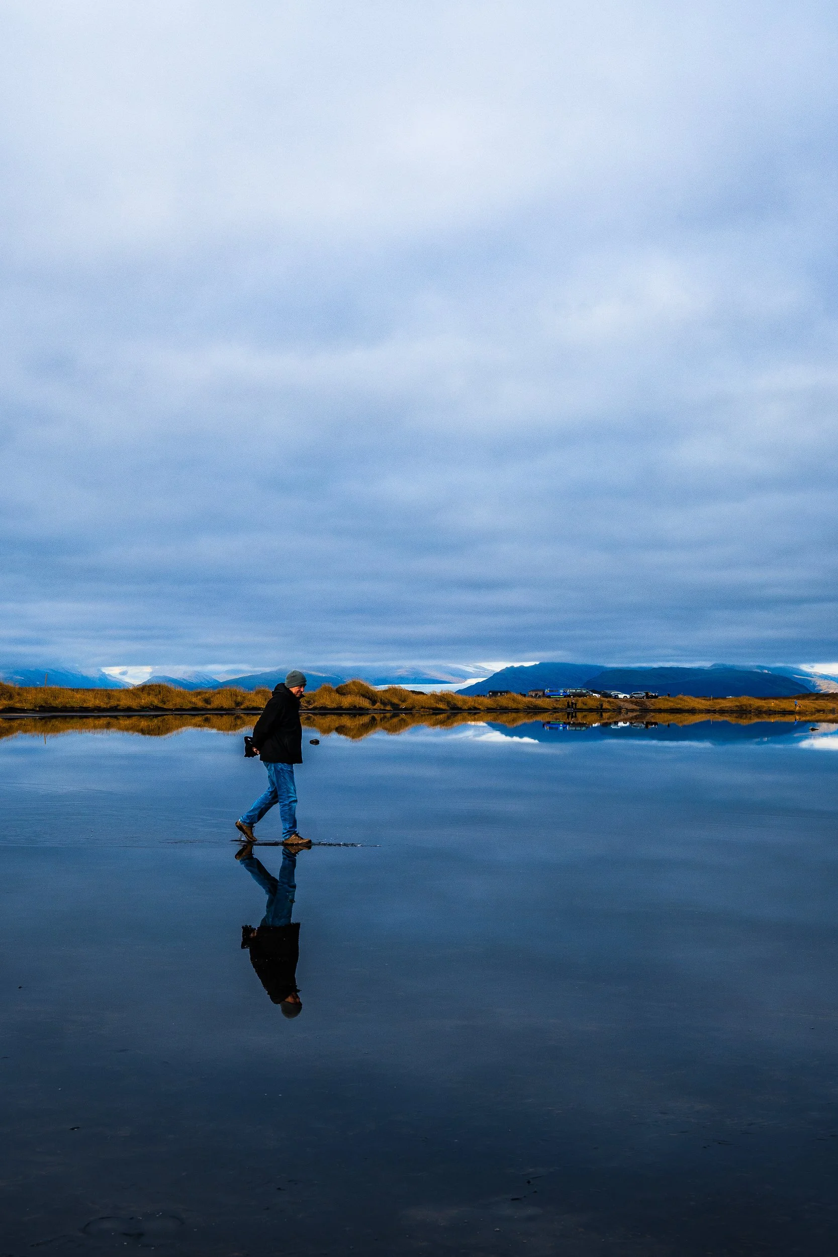 Person walking across a reflective lake with snow-capped mountains and cloudy blue sky in the background