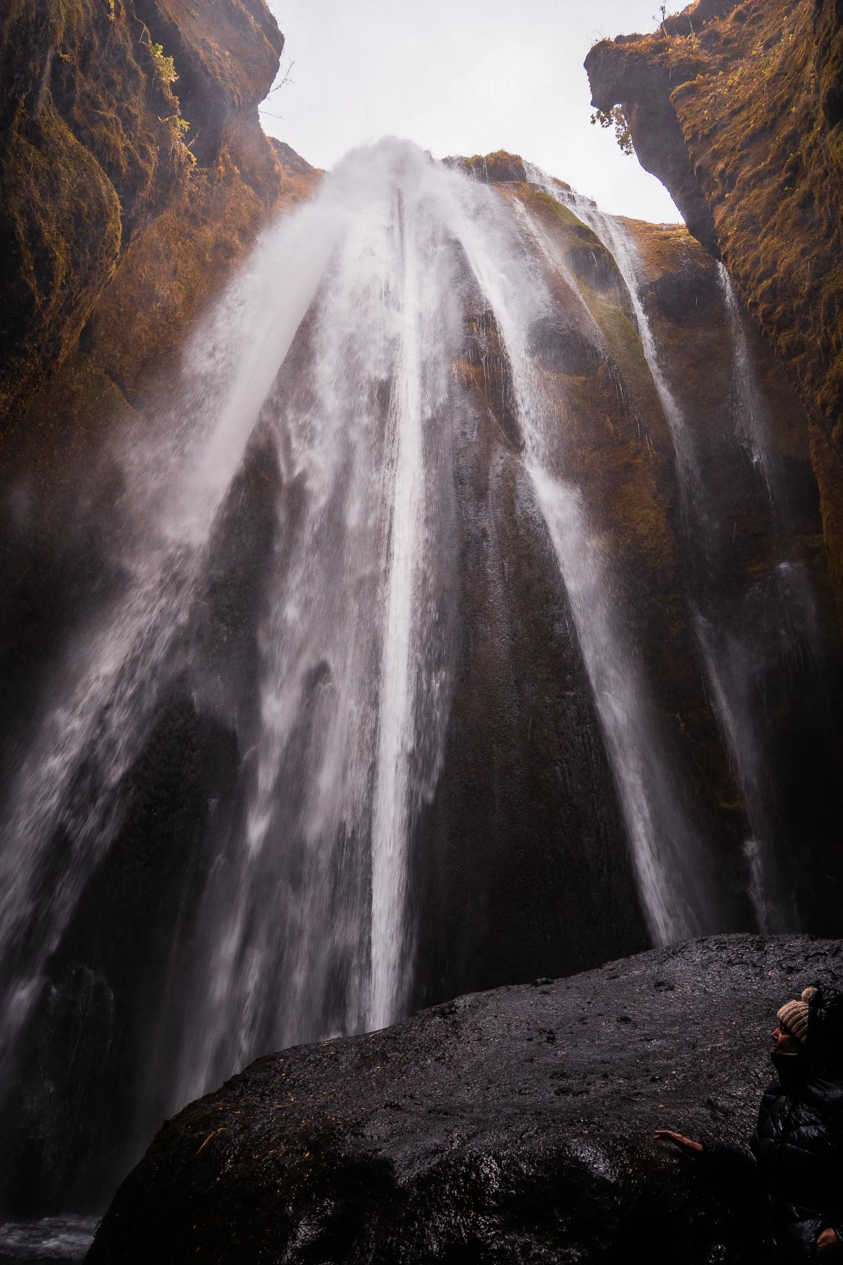 Waterfall cascading over moss-covered cliffs, viewed from inside a rocky canyon with a person in the foreground
