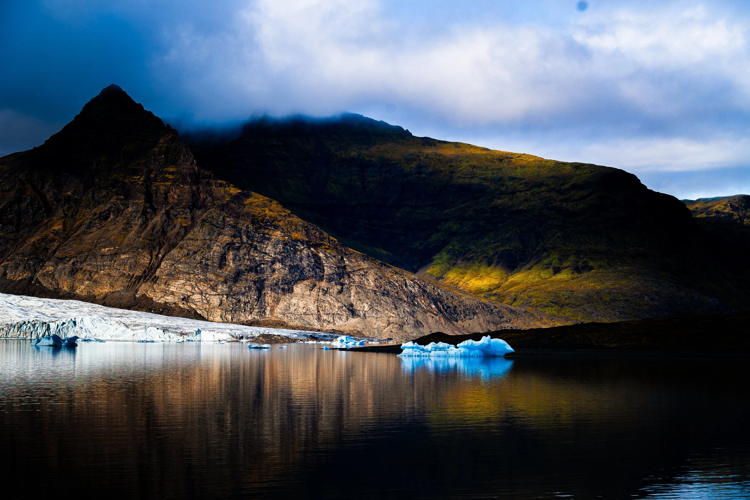 Mountain glacier meeting a calm lake with floating blue icebergs under cloudy skies