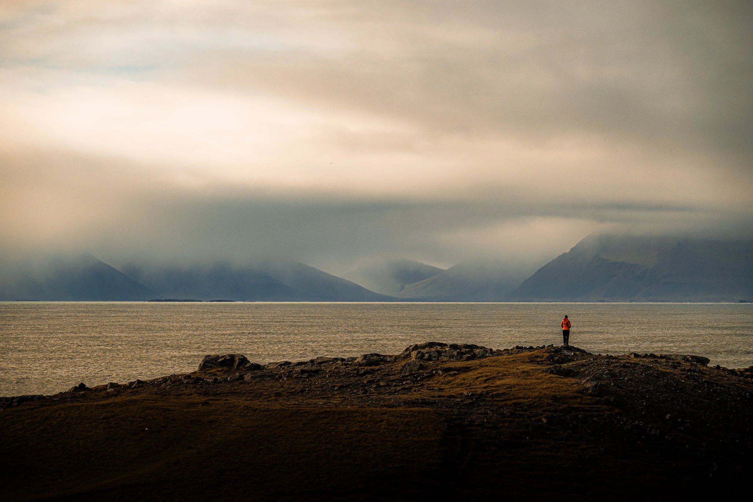 Person in an orange jacket standing on a rocky shoreline overlooking a calm sea and mist-covered mountains.