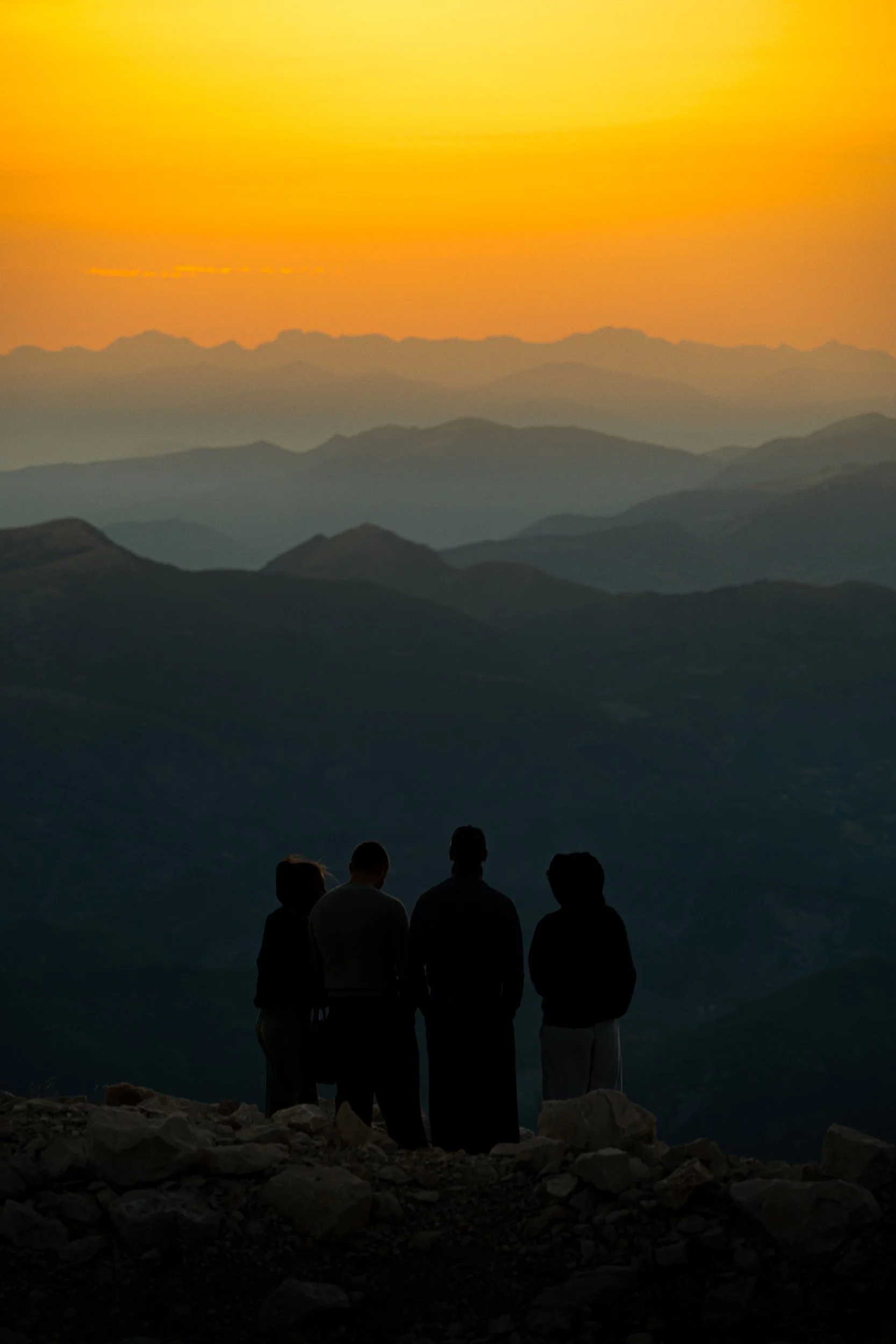 Four people silhouetted on a rocky mountain overlook at sunset, facing layered peaks beneath an orange sky.