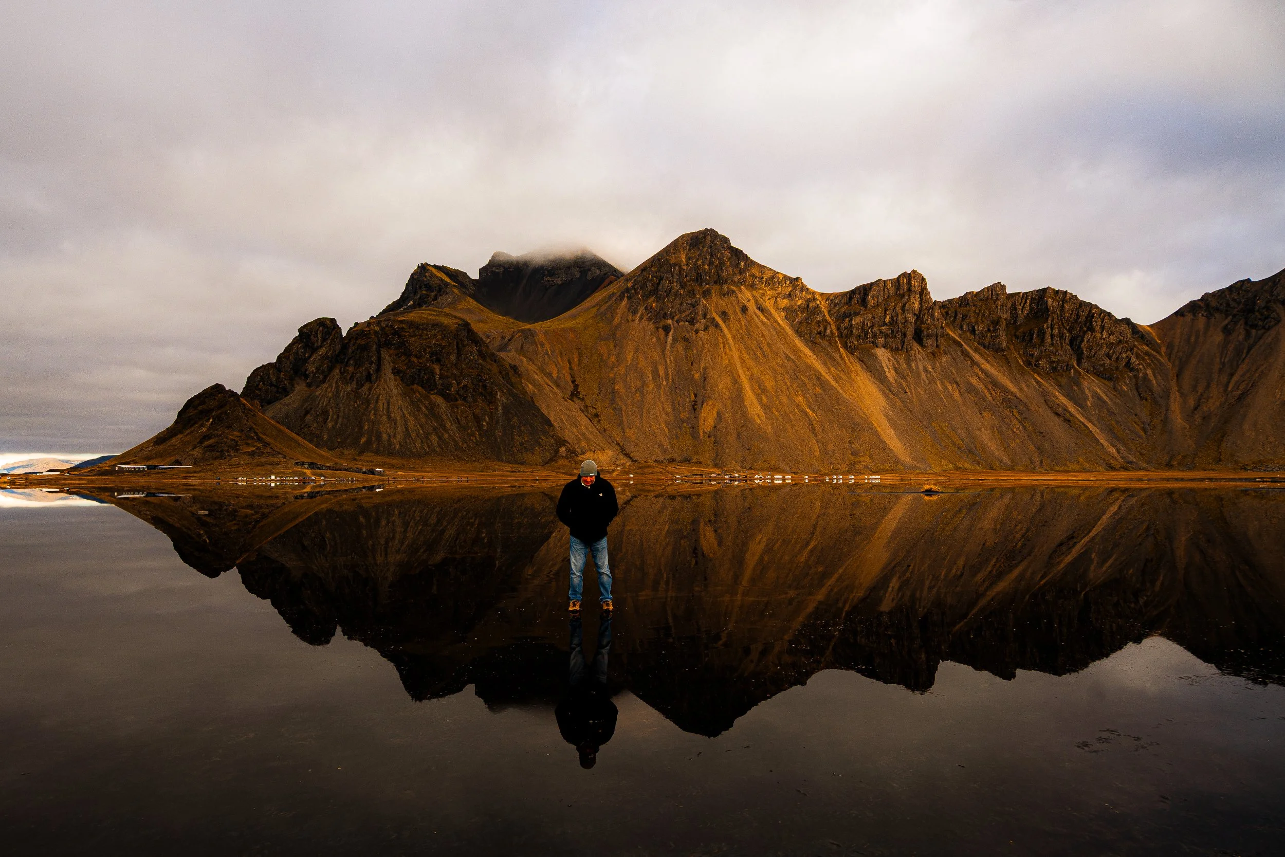 Person standing beside a calm lake reflecting rugged golden mountain peaks under a cloudy sky