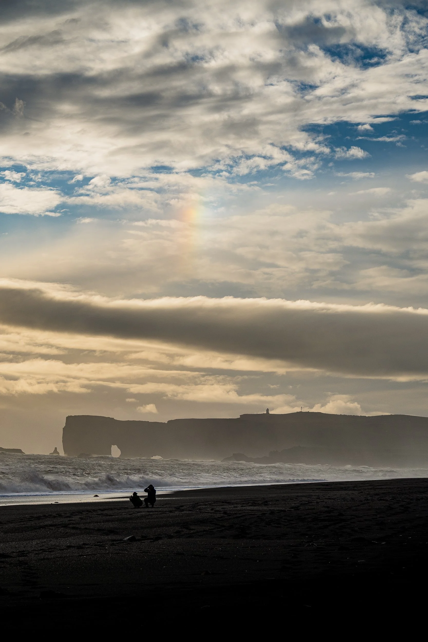 Silhouetted people on a black sand beach beneath dramatic clouds, ocean waves, and a distant sea cliff with a natural arch.
