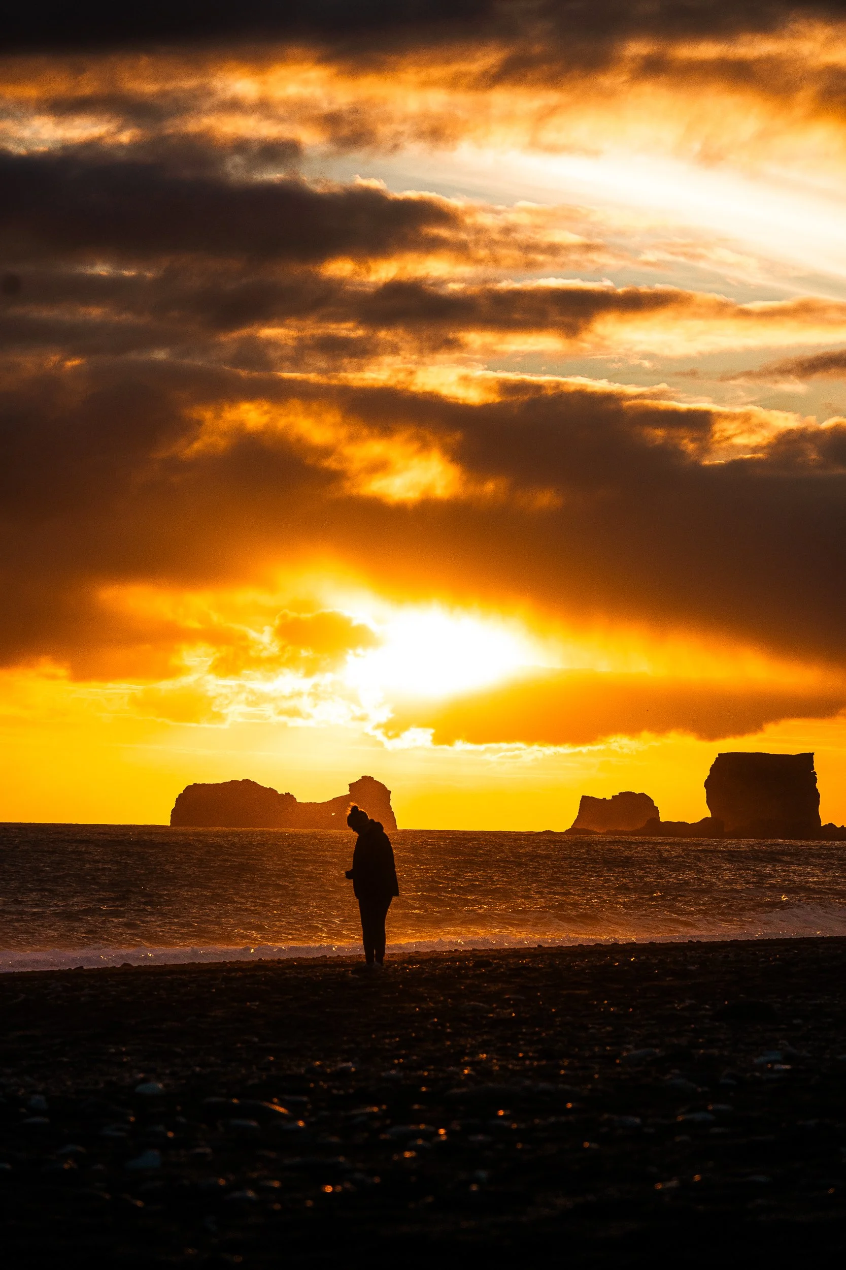Silhouetted person standing on a beach at sunset beneath dramatic golden clouds and offshore rock formations