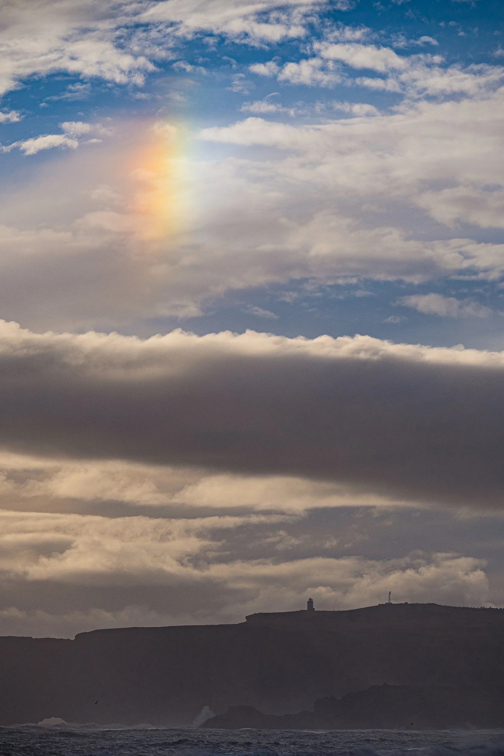Dramatic ocean coastline with a dark cliff silhouette, lighthouse, crashing waves, layered storm clouds, and a faint rainbow.