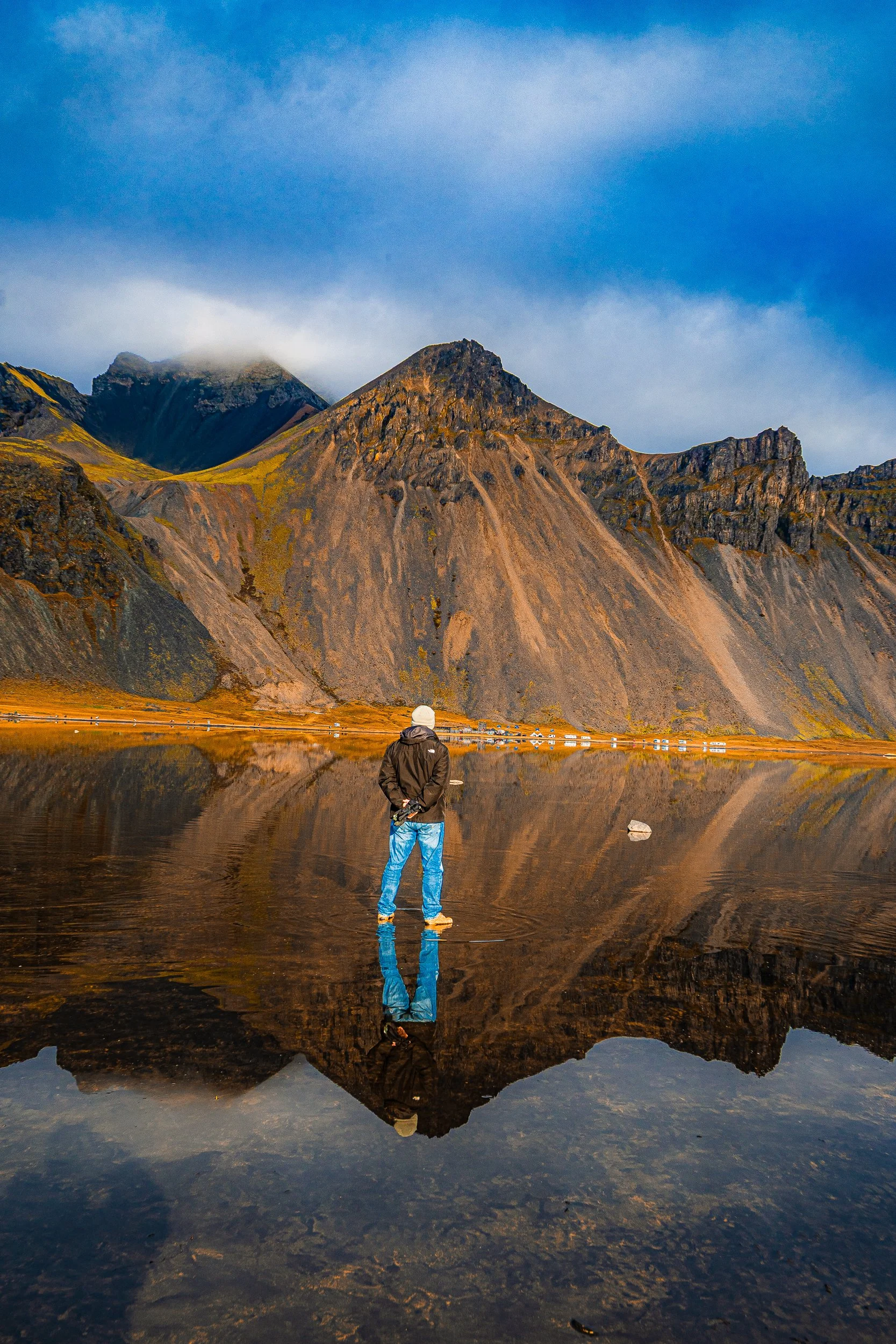 Person standing in a shallow reflective lake before rugged, sunlit mountains beneath a cloudy blue sky