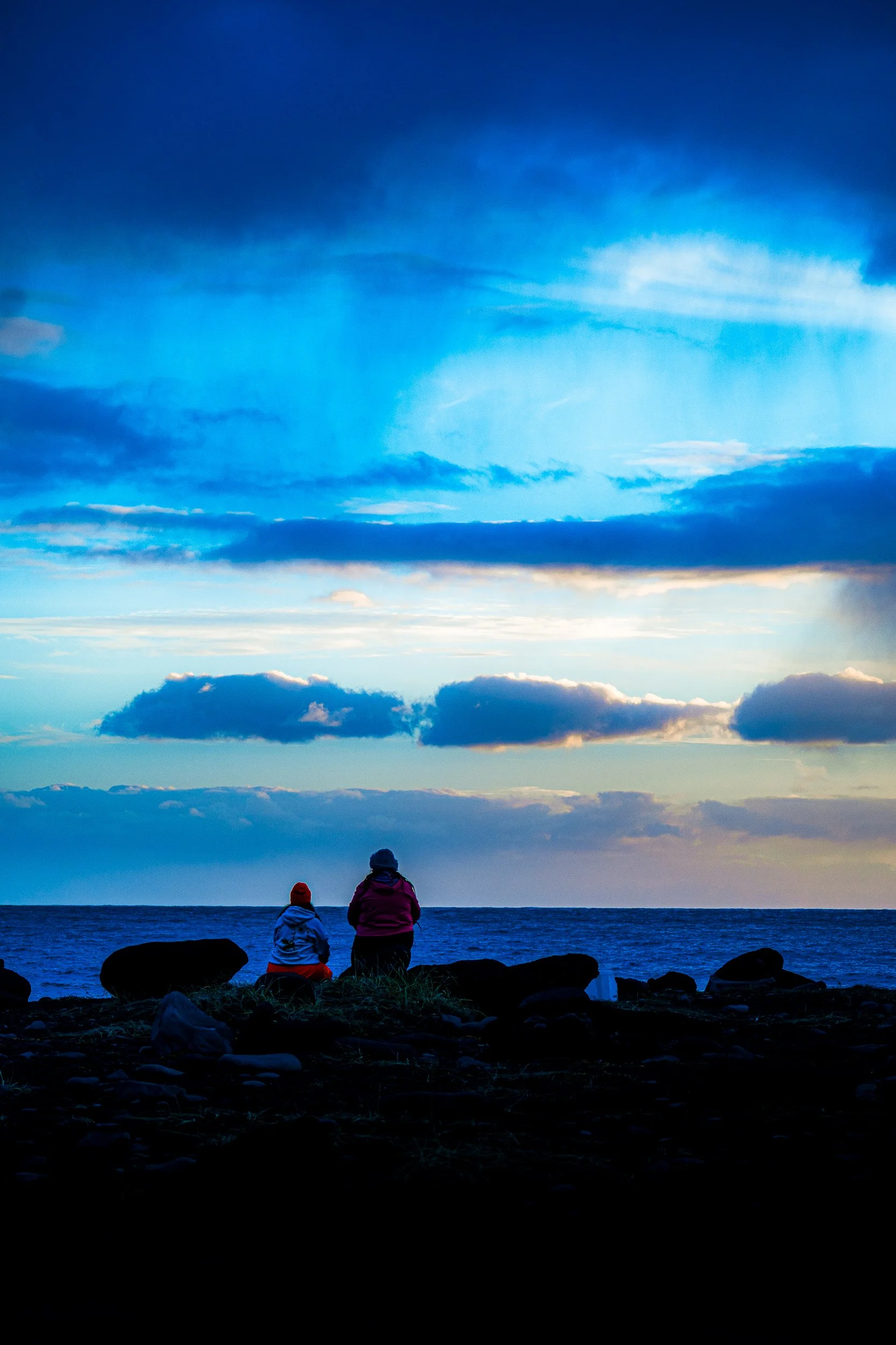Two people sitting on a rocky shoreline, watching a cloud-filled sunset over the ocean.