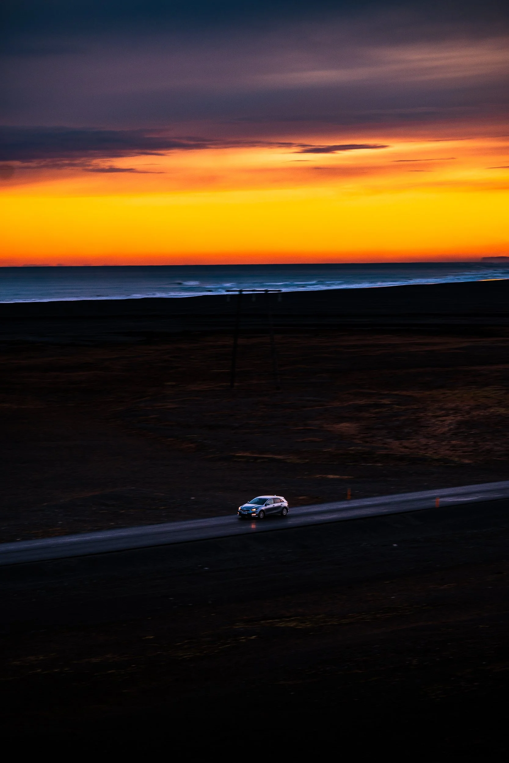 White hatchback driving along a coastal road beside the ocean at sunset