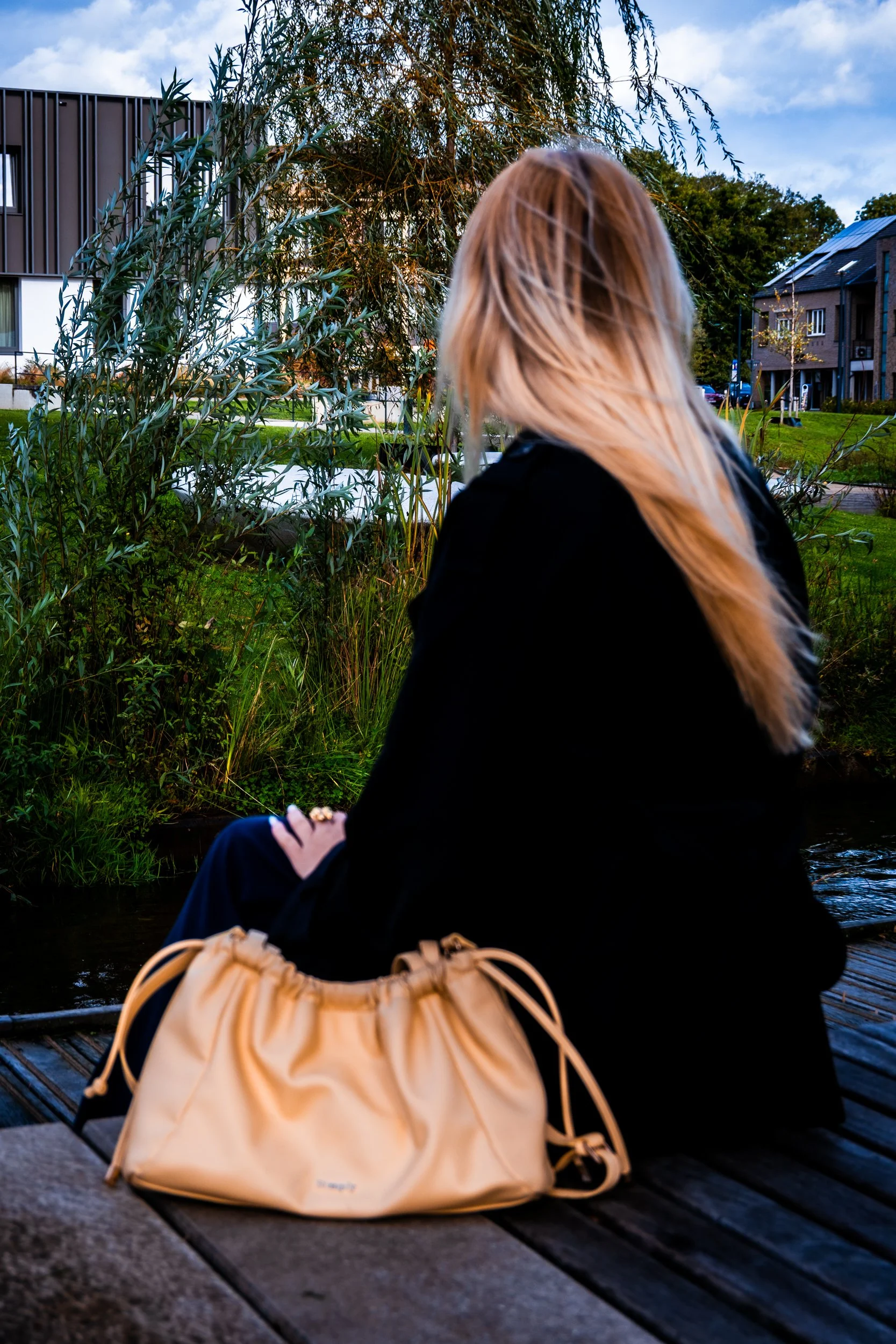 Woman with long blonde hair sitting by a canal with a beige drawstring bag, greenery, and buildings in the background