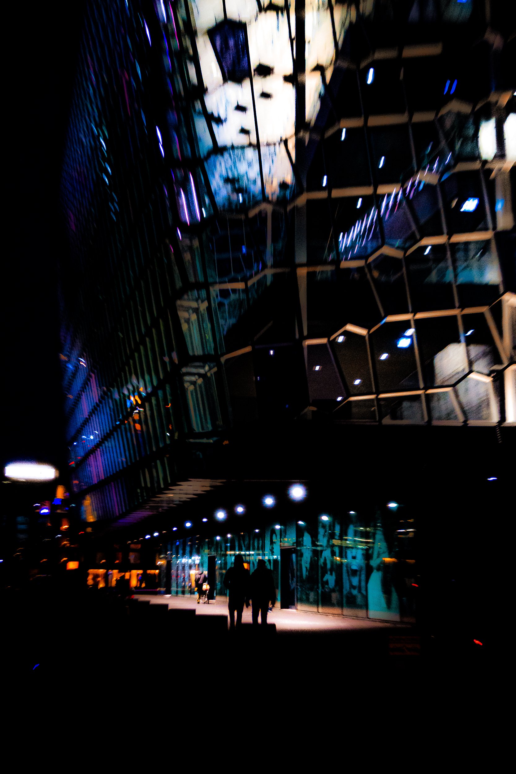 Abstract nighttime cityscape with illuminated geometric building reflections, streetlights, and silhouetted pedestrians