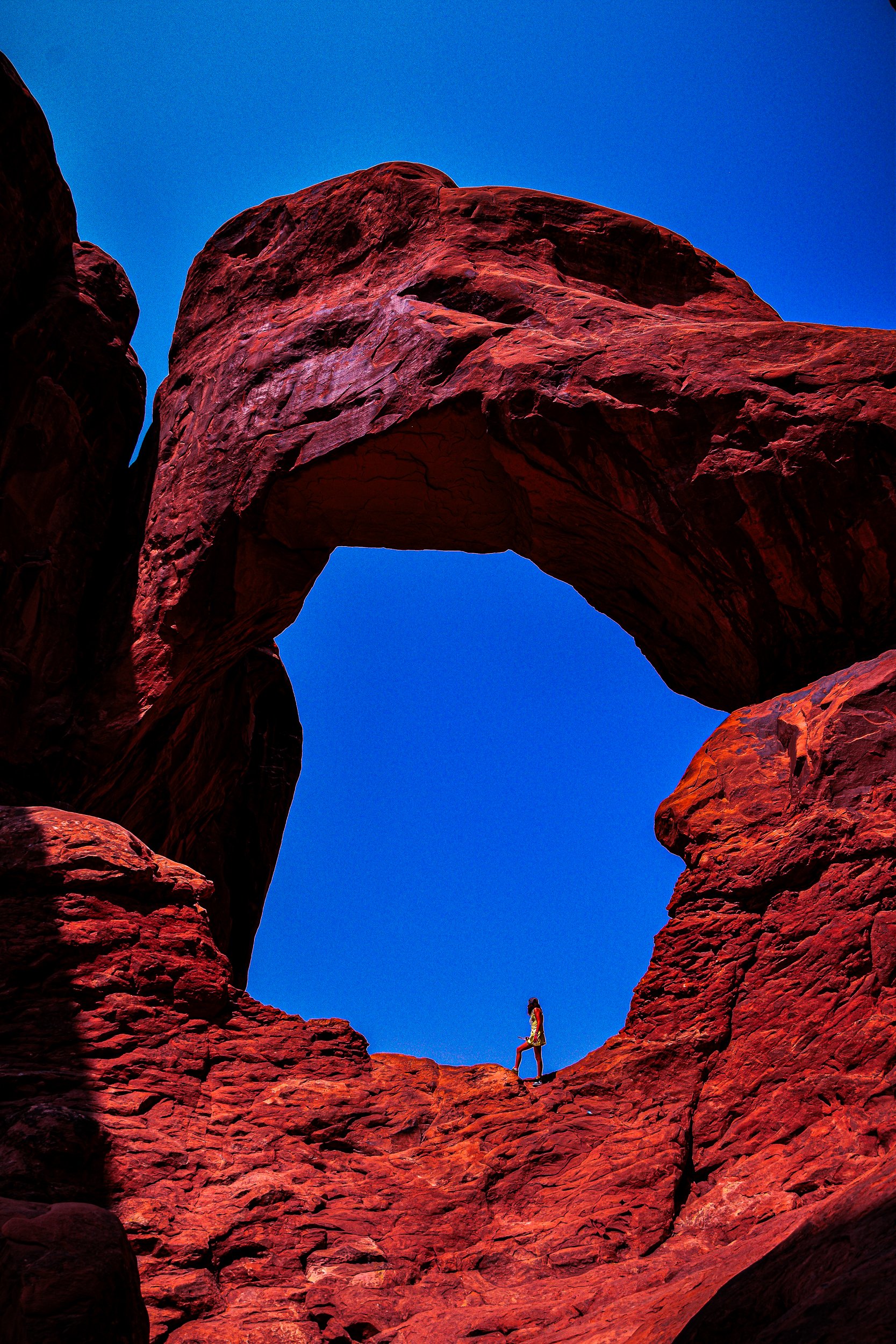 Hiker standing beneath a massive red sandstone arch against a vivid blue sky