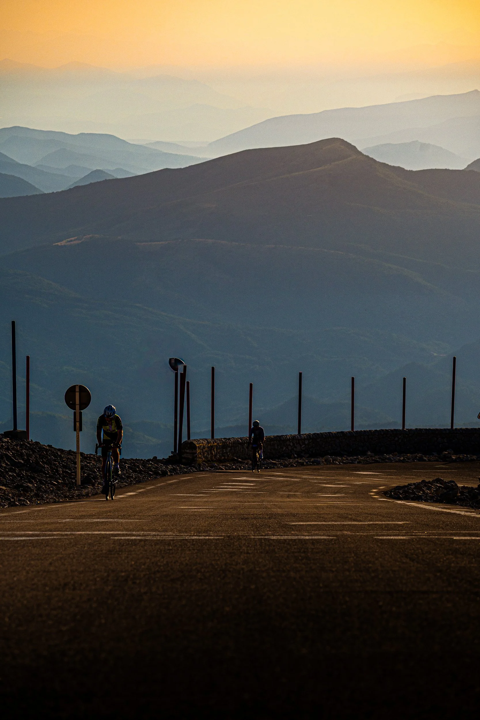 Cyclists climbing a mountain road with layered peaks in the background at sunset
