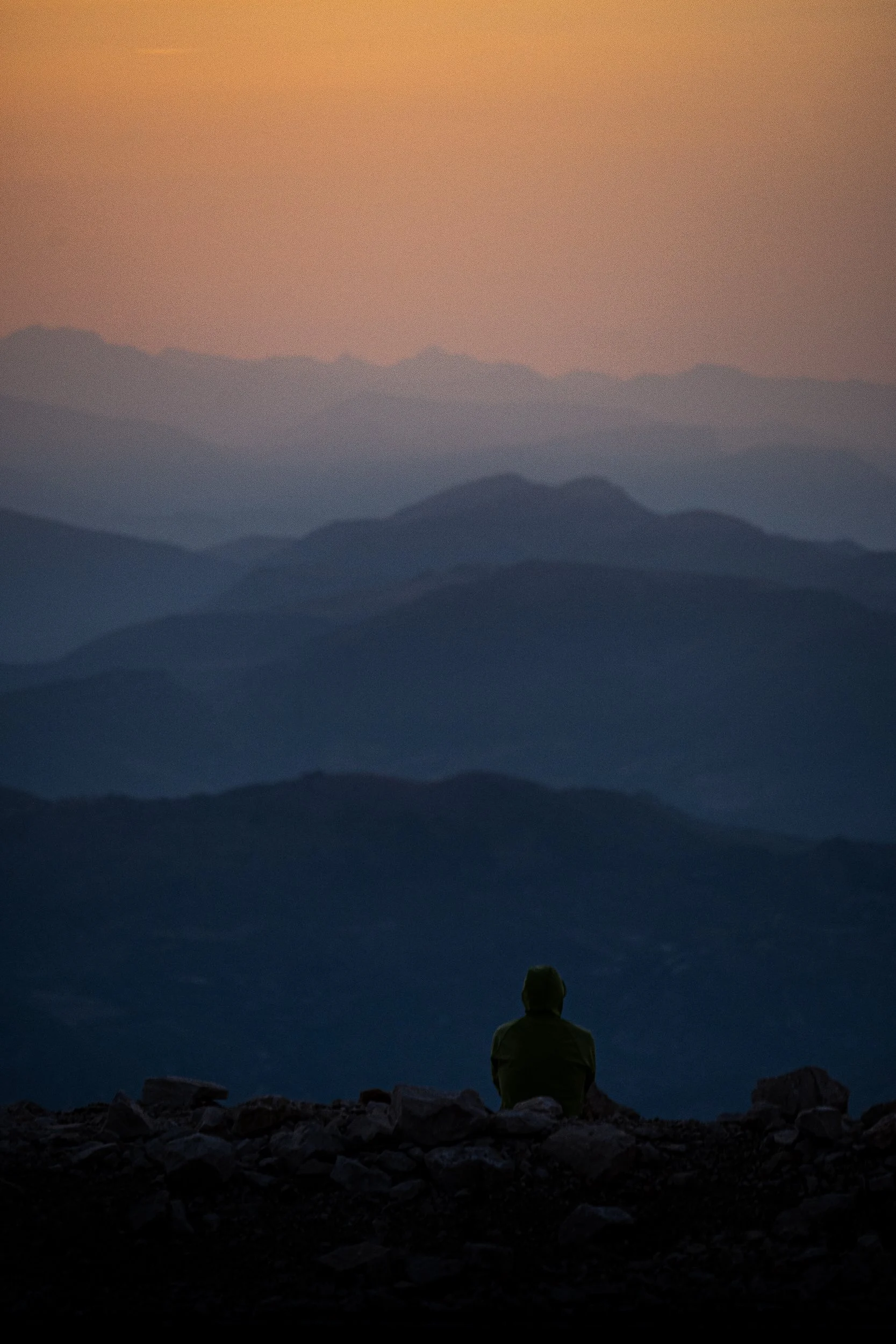 Silhouetted person sitting on rocky terrain overlooking layered mountain ranges at sunset
