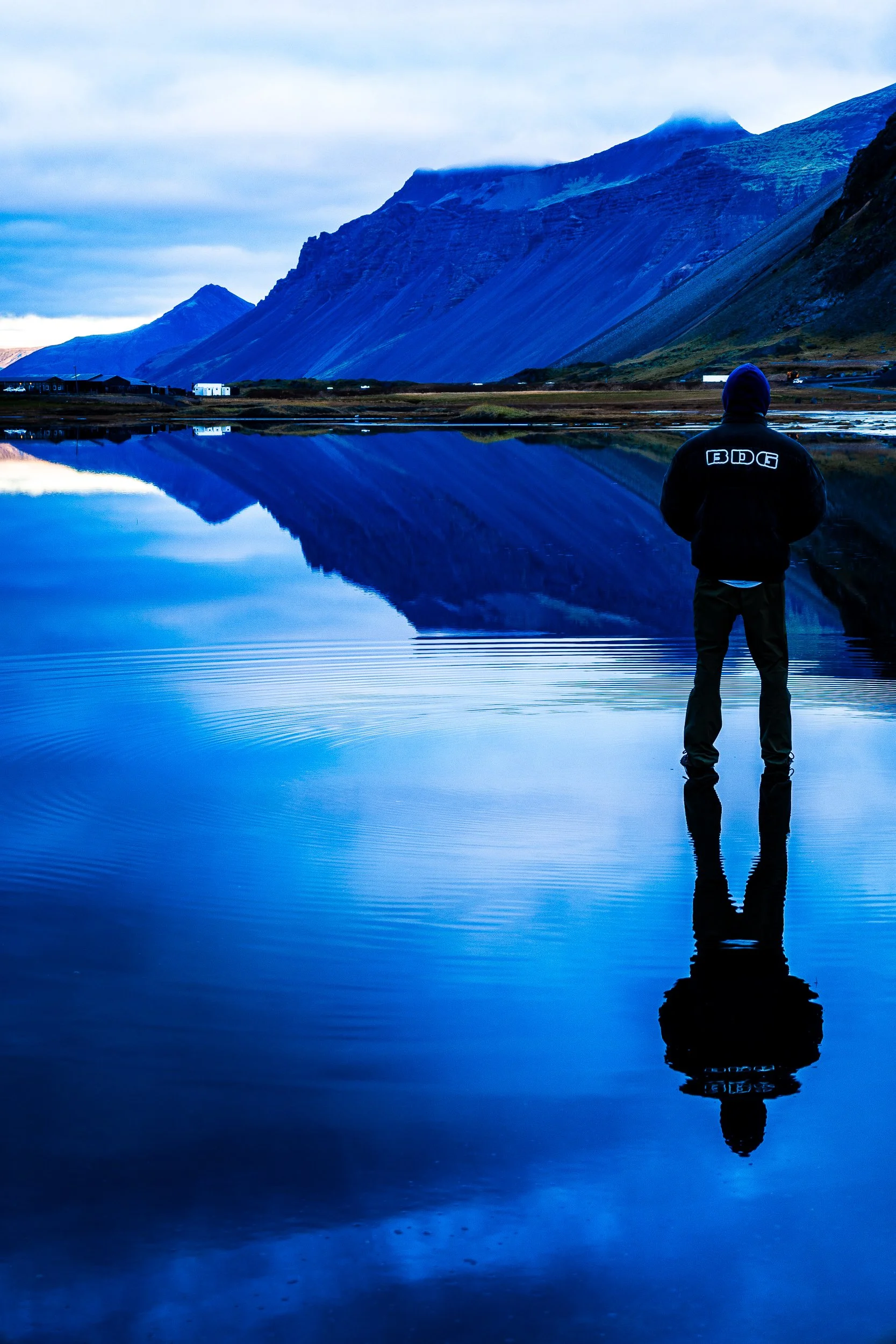 Person in a hooded jacket standing beside a calm lake reflecting dramatic blue mountains and cloudy skies