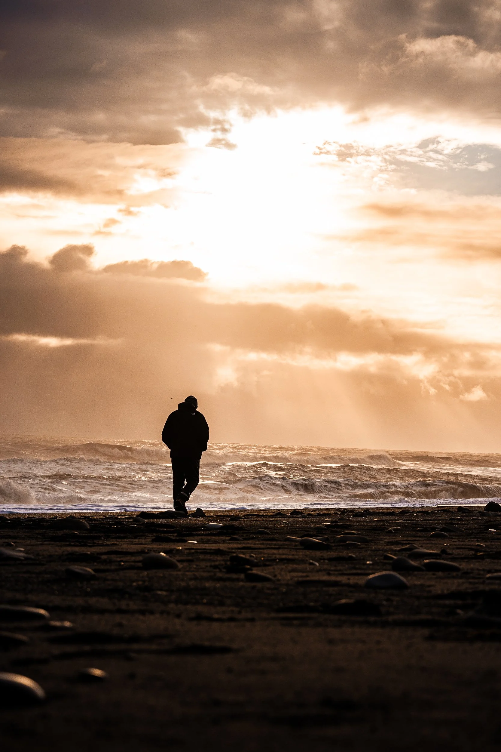 Silhouetted person walking along a rocky beach beside rough ocean waves at sunset