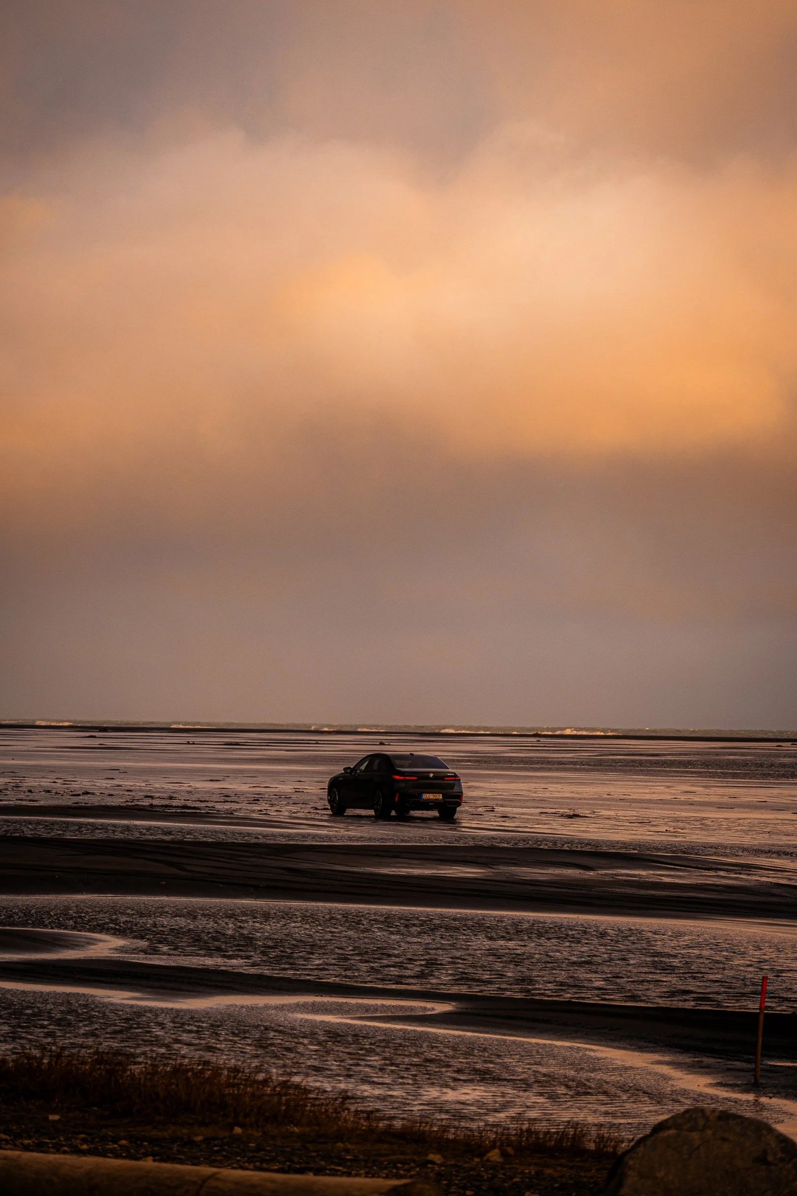 Black car driving across a tidal beach at sunset under a hazy orange sky