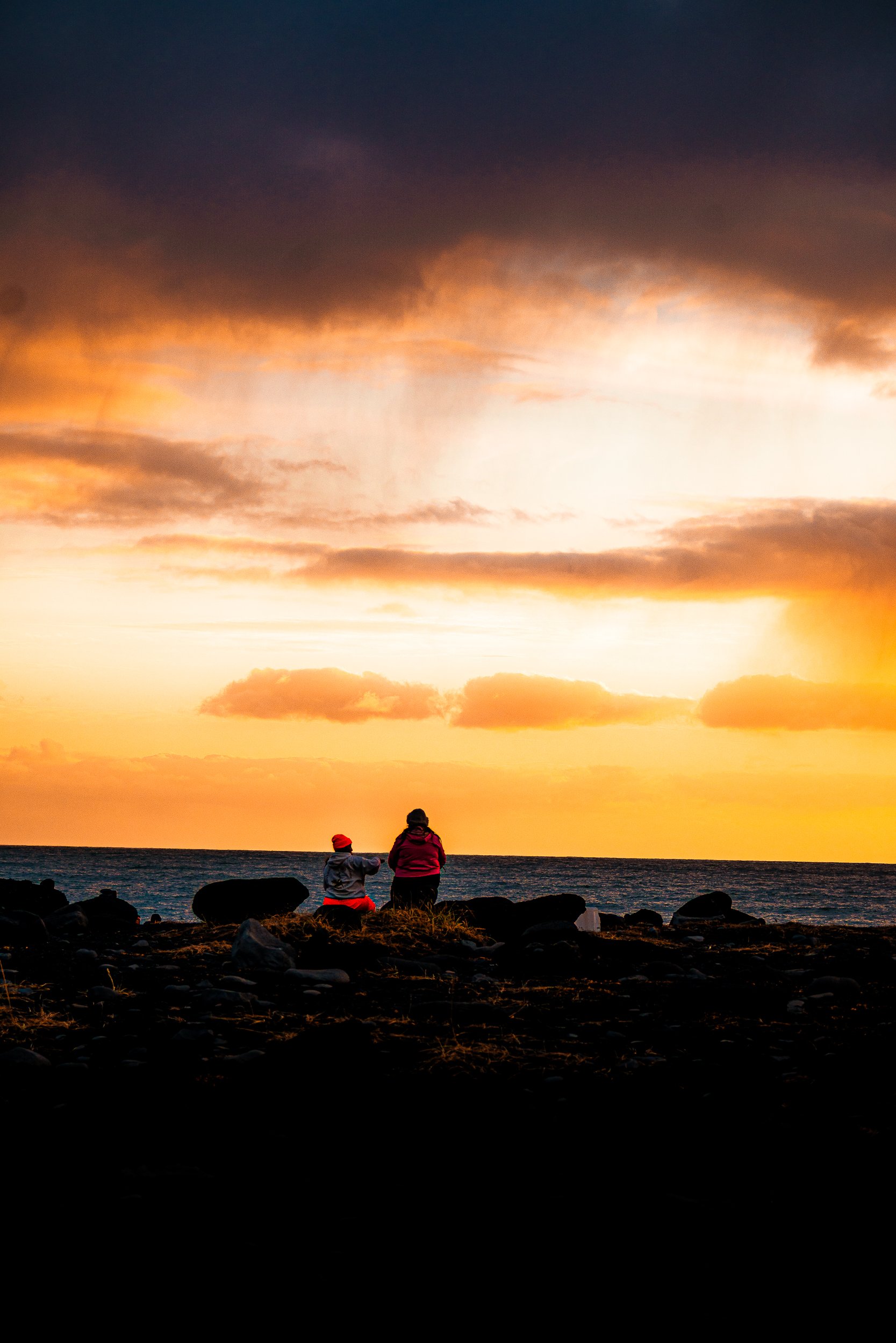 Two people sitting on a rocky shoreline overlooking the ocean at golden sunset