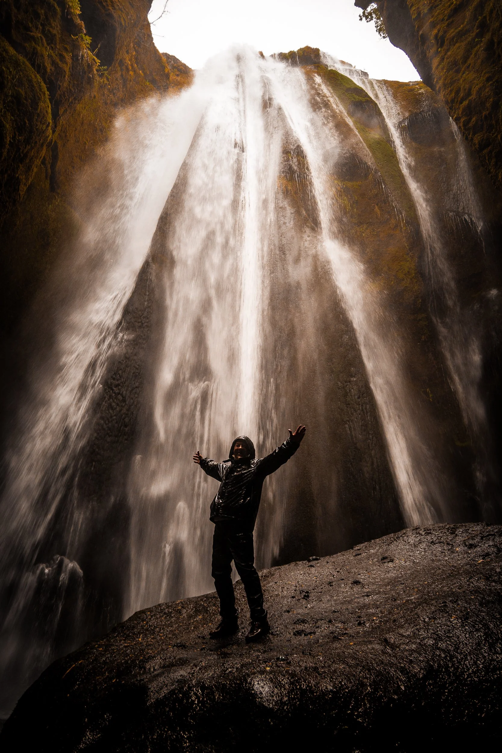 Person in a black rain jacket standing with arms outstretched beneath a towering waterfall in a moss-covered canyon