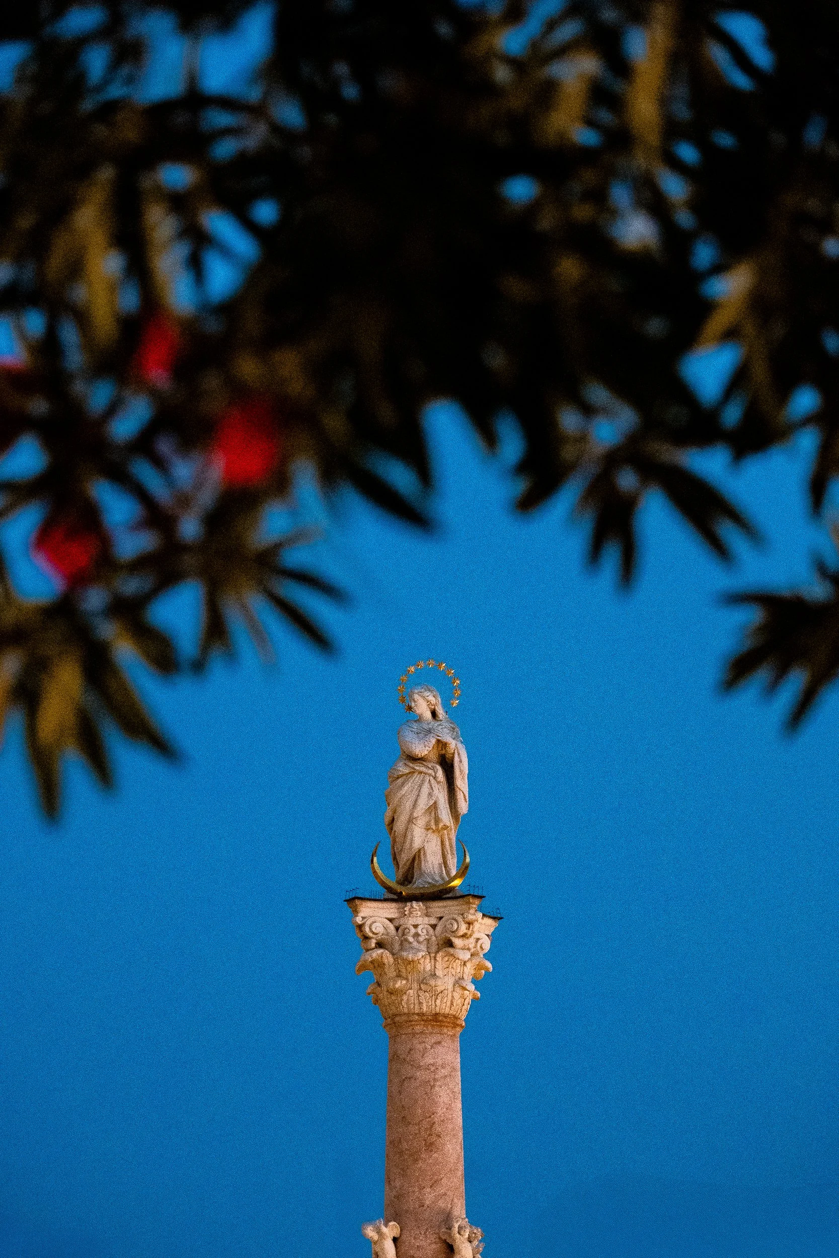 Statue of the Virgin Mary atop an ornate column against a vivid blue sky, framed by blurred tree branches.