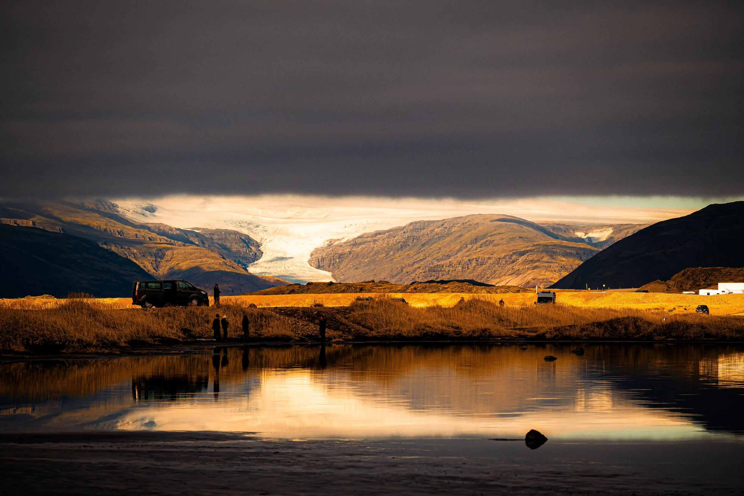 Glacier-fed mountain valley reflected in a lake beneath dark storm clouds, with visitors and vehicles in the foreground