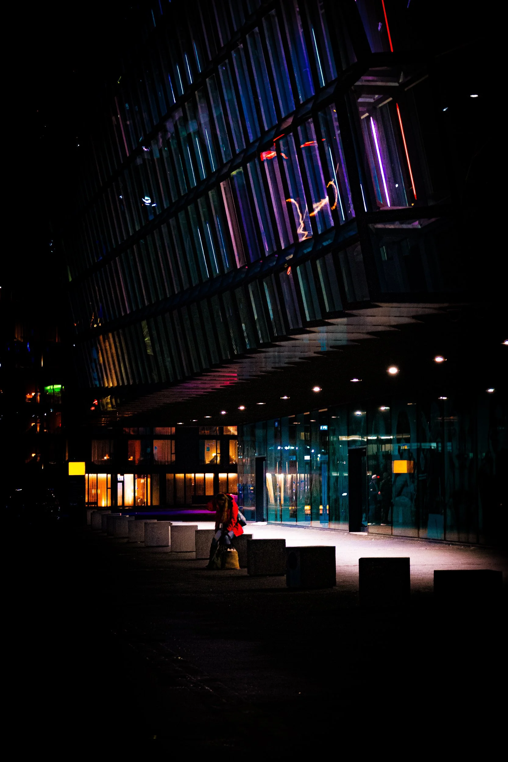 Modern glass building at night illuminated by colorful neon lights, with a seated person outside.