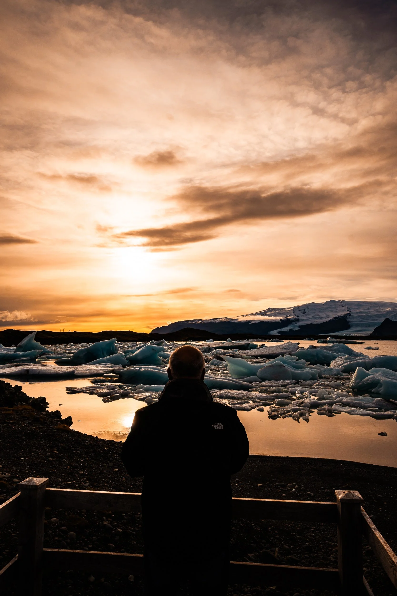 Person viewing floating icebergs on a glacial lagoon beneath a snowy mountain at sunset