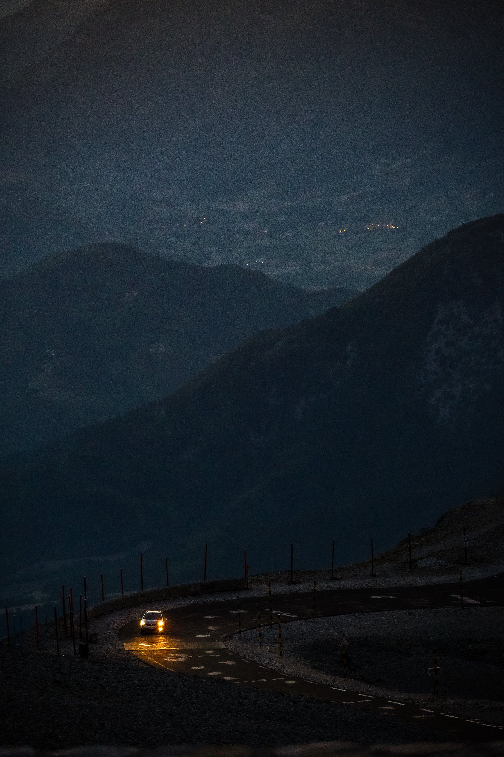 Car with headlights on driving along a wet mountain road at night, surrounded by misty dark mountain peaks and distant lights