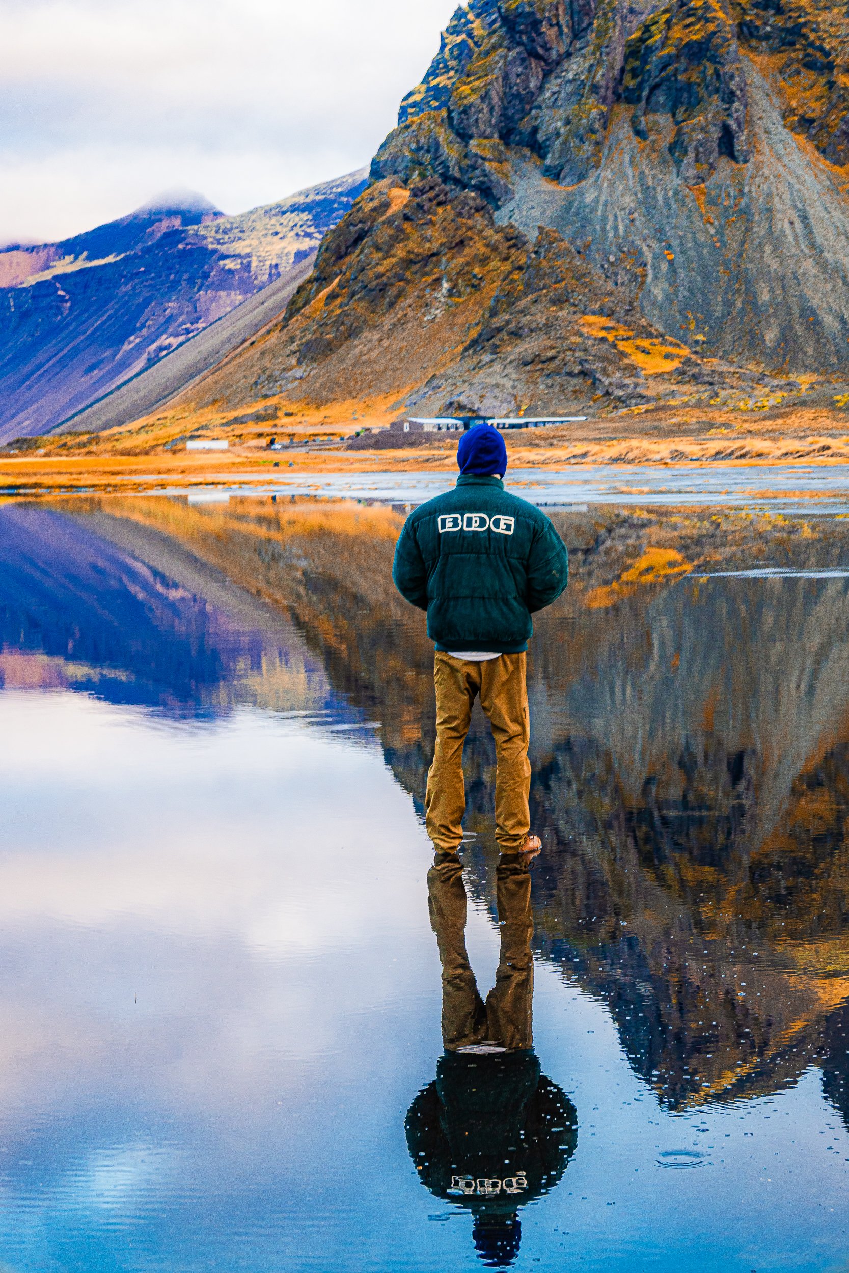 Person standing in shallow mountain lake, reflected beneath towering rocky cliffs and autumn-colored peaks