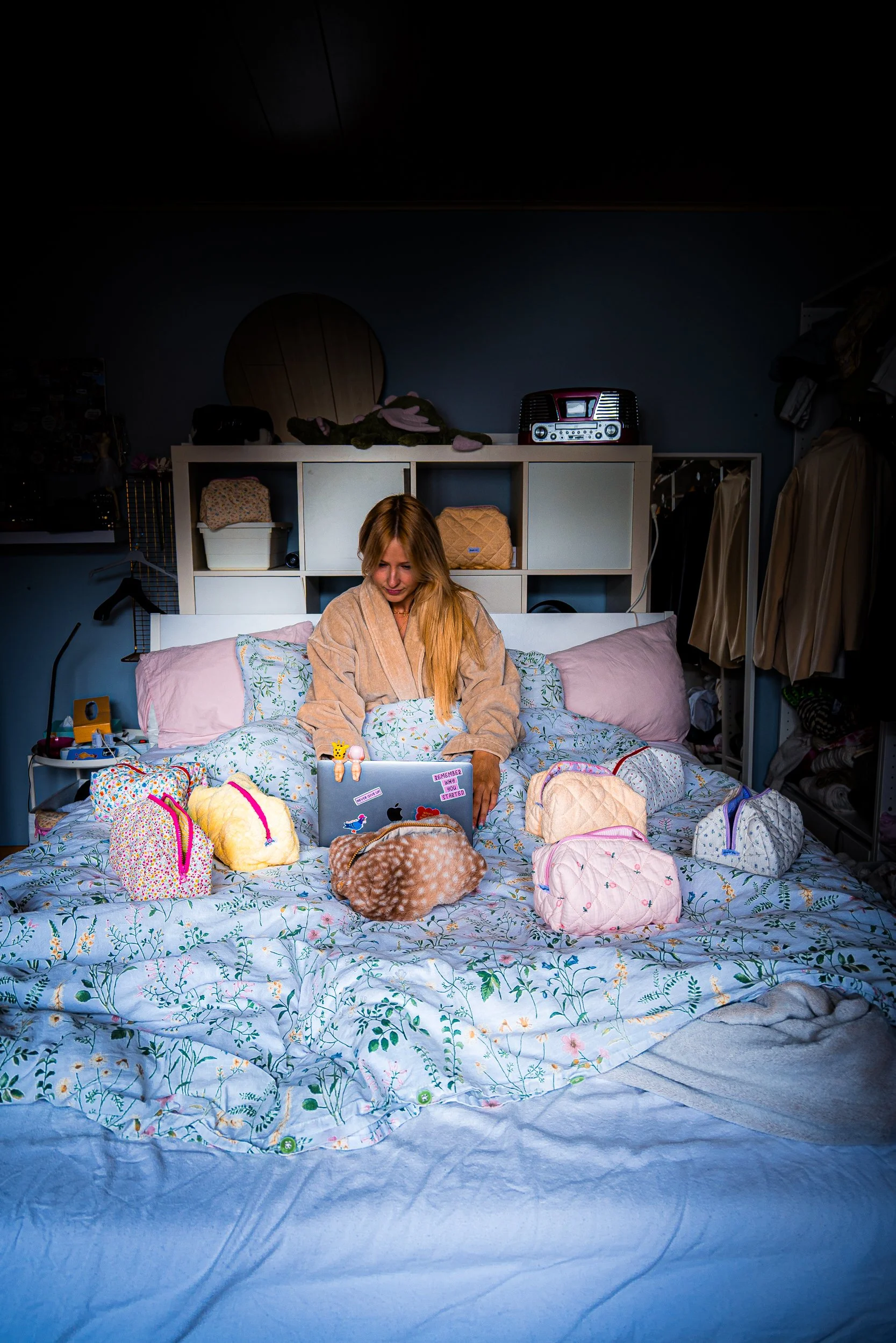 Woman in a robe using a sticker-covered laptop on a floral bed in a cozy bedroom
