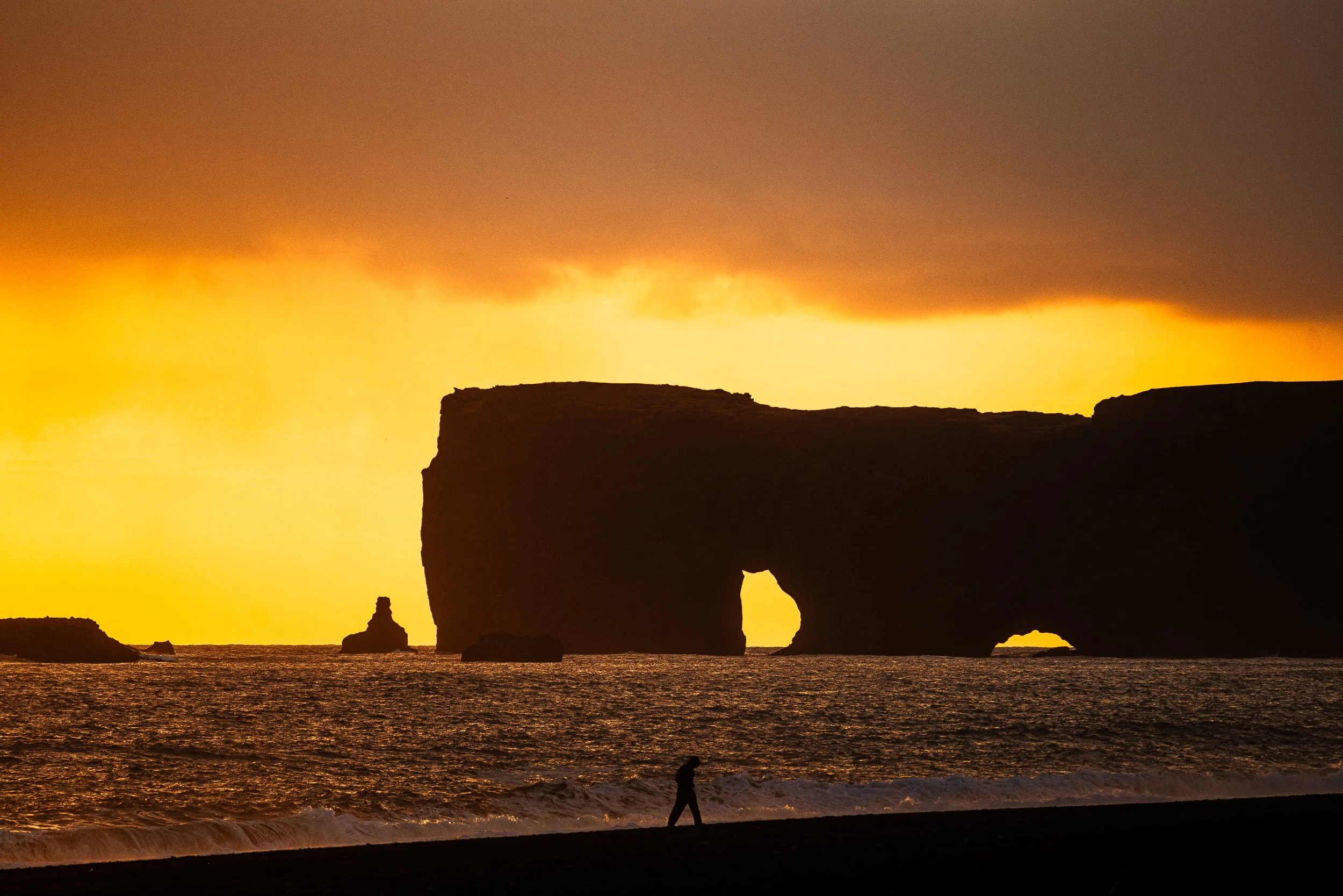 Silhouette of Reynisdrangar sea stacks and a person walking along a beach at sunset in Iceland
