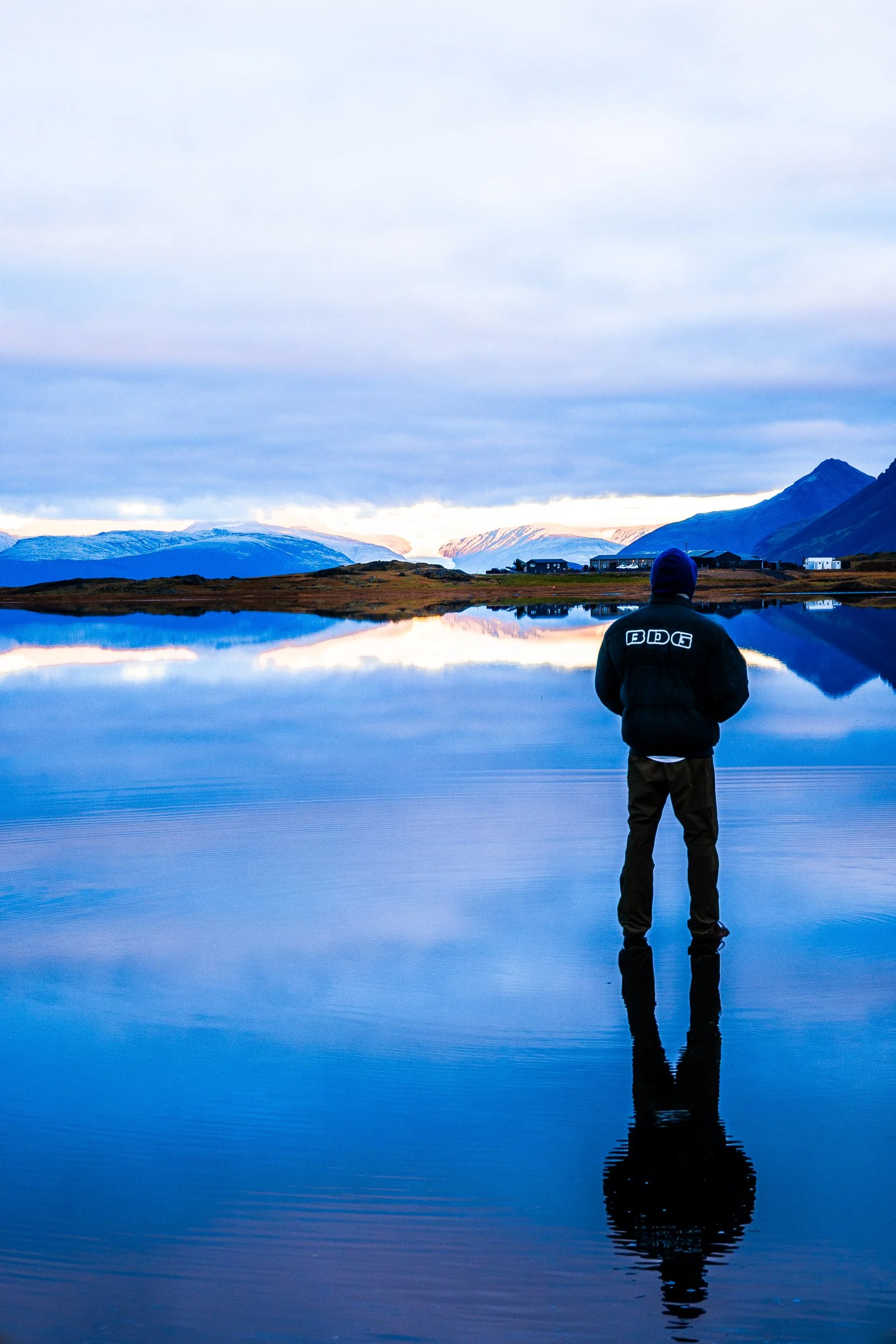 Person in a hooded jacket standing on a reflective lake in front of snow-capped mountains and a mountain settlement