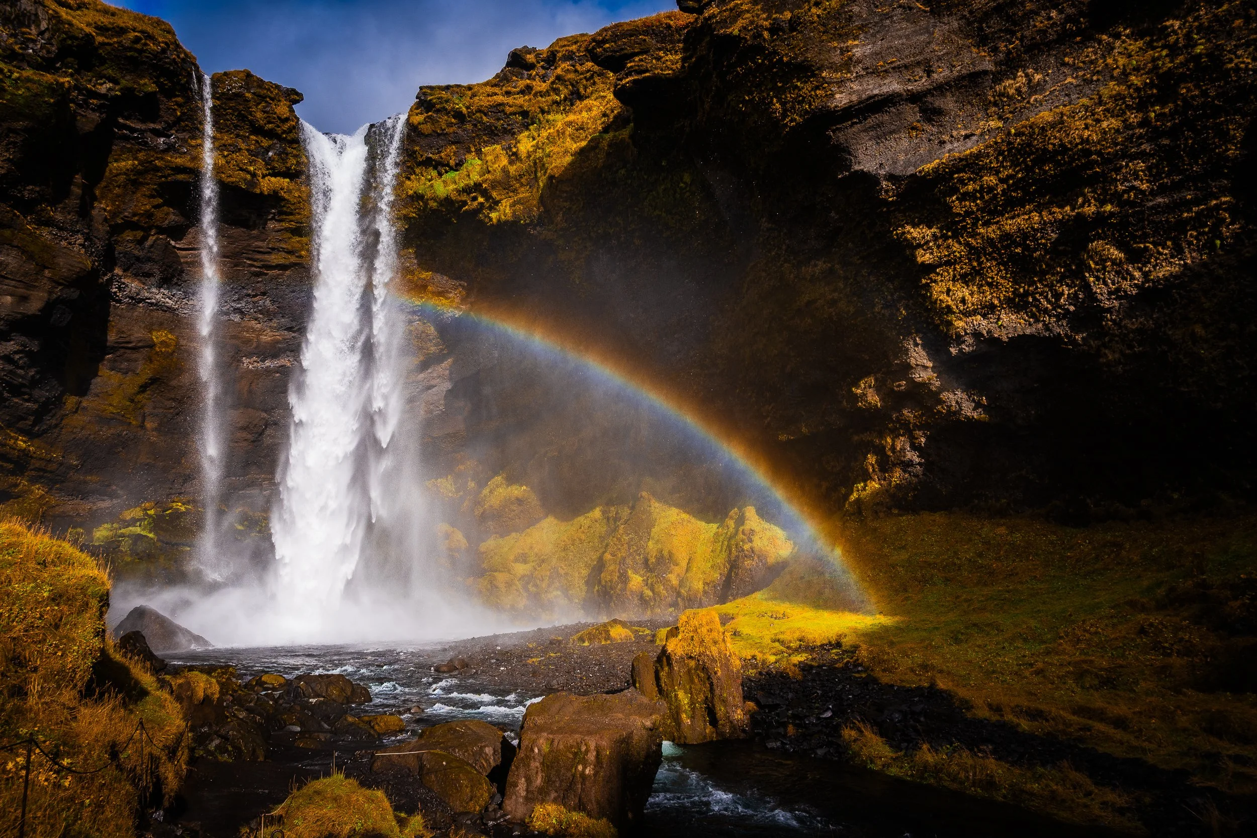 Waterfall cascading over moss-covered cliffs with a rainbow in the mist and a rocky stream below