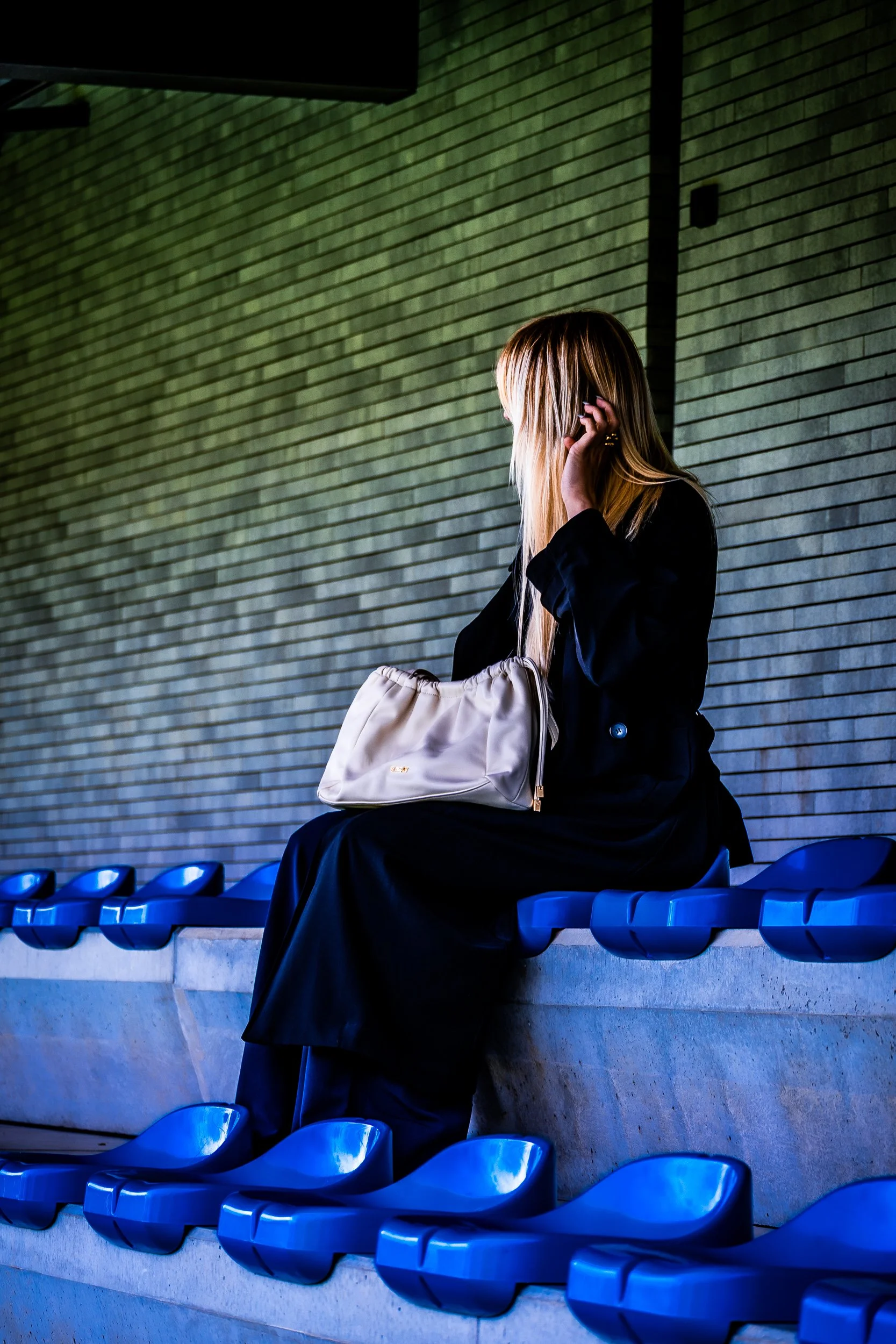 Woman in a black coat sitting alone in blue stadium seats with a white handbag.