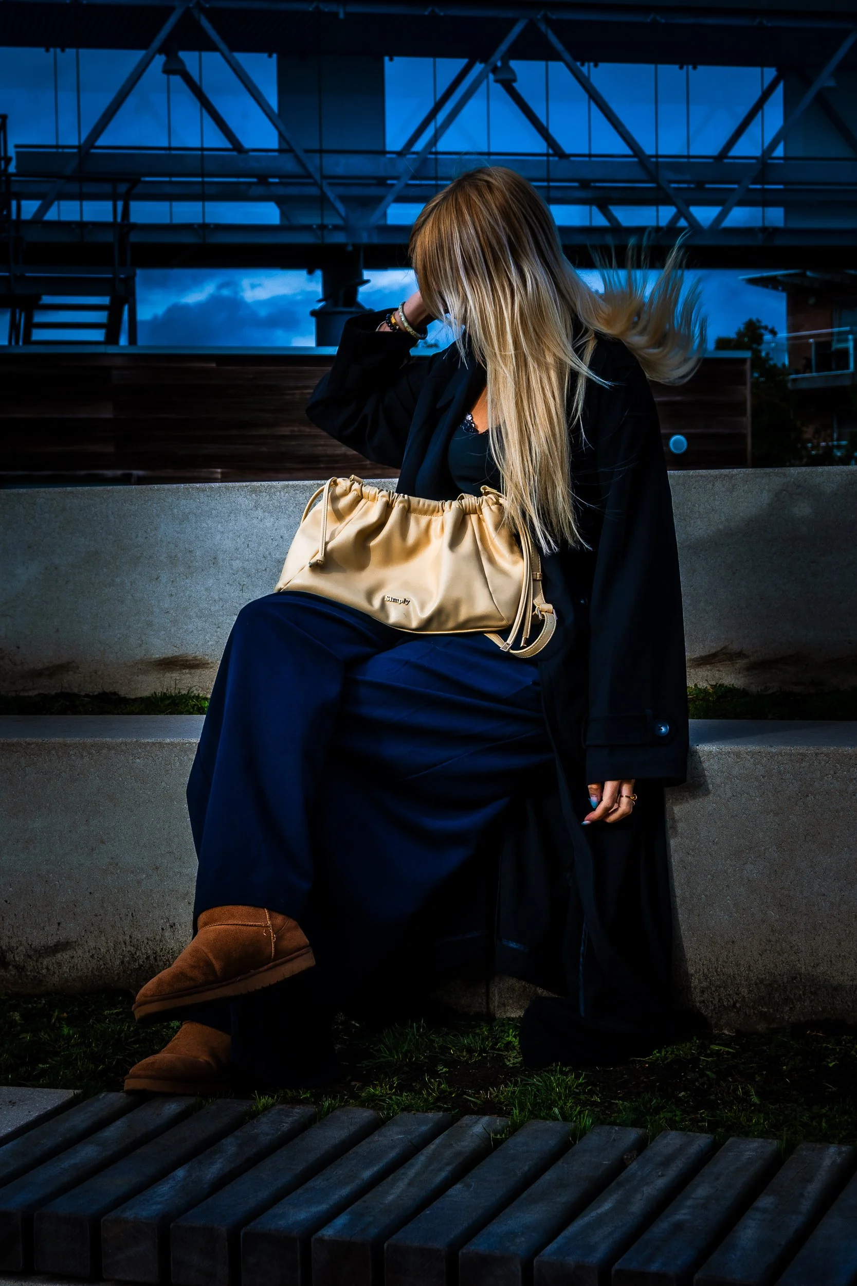 Woman with long blonde hair sitting outdoors in a black coat and navy pants, holding a tan handbag.