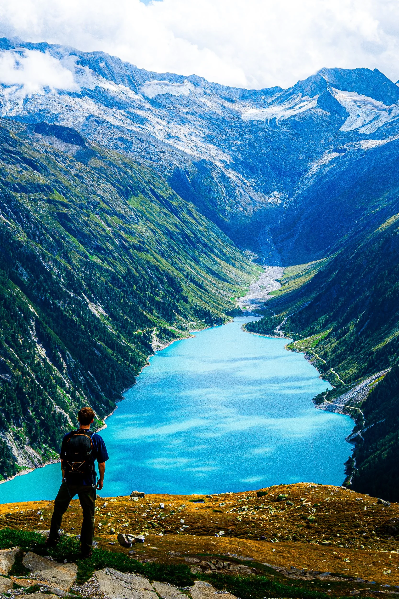 Hiker with a backpack overlooking a turquoise alpine lake, glacial valley, and snow-capped mountains