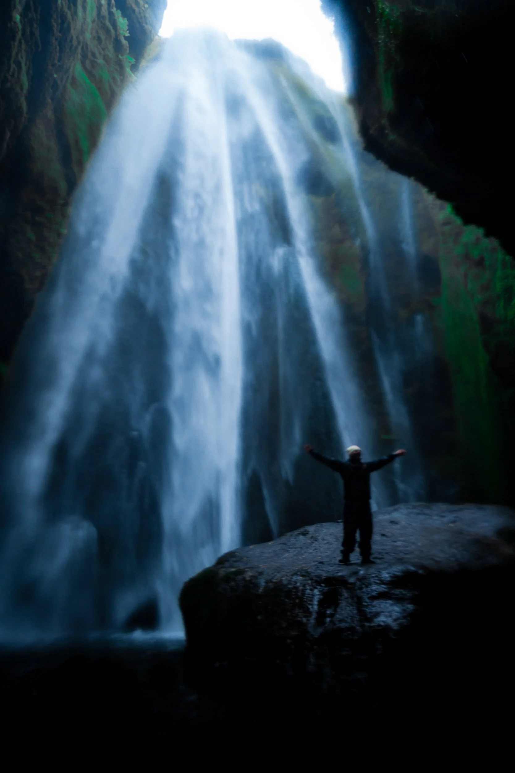 Person with arms outstretched in front of a towering waterfall surrounded by forest