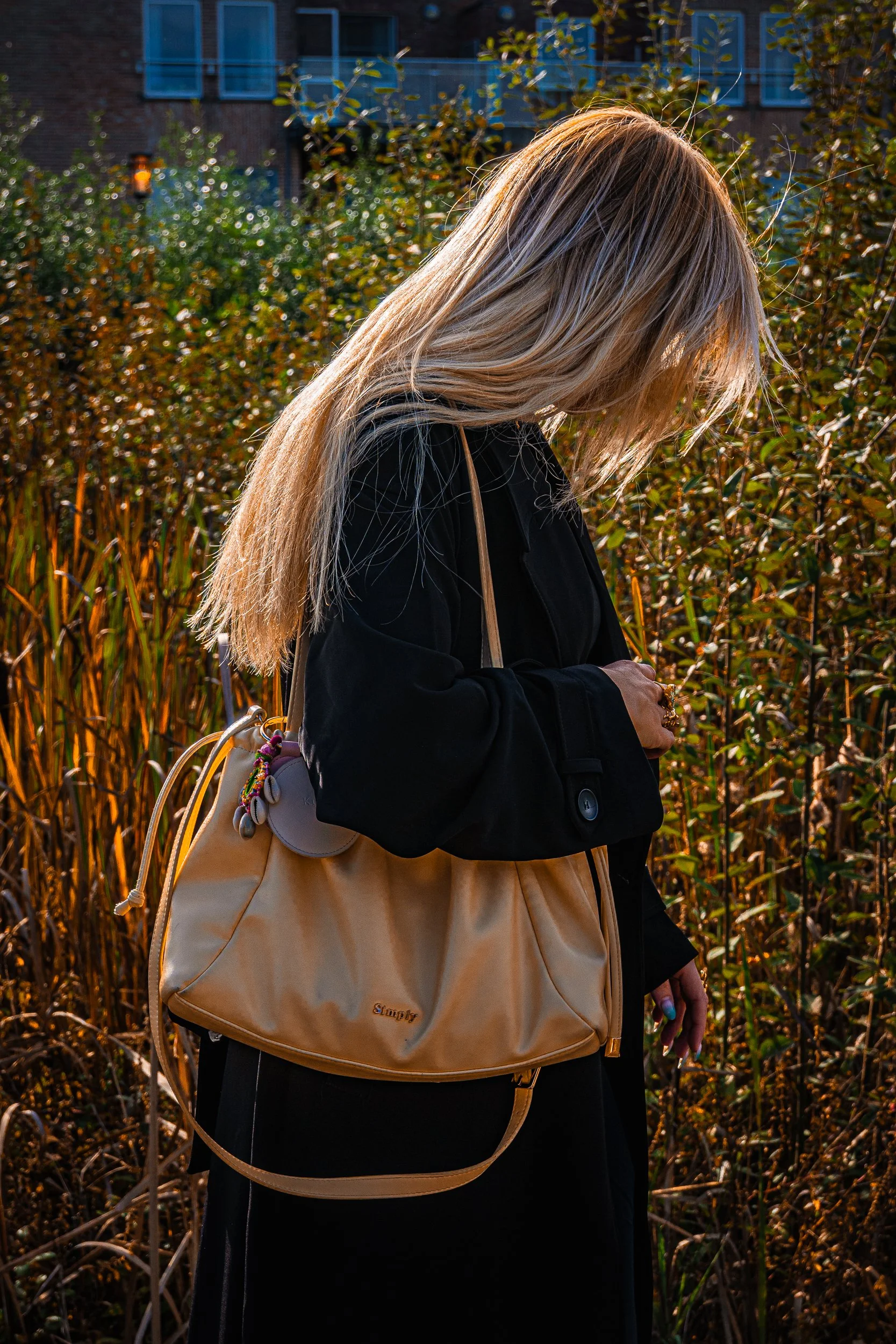 Person with long hair wearing a black coat and carrying a tan shoulder bag in a sunlit garden
