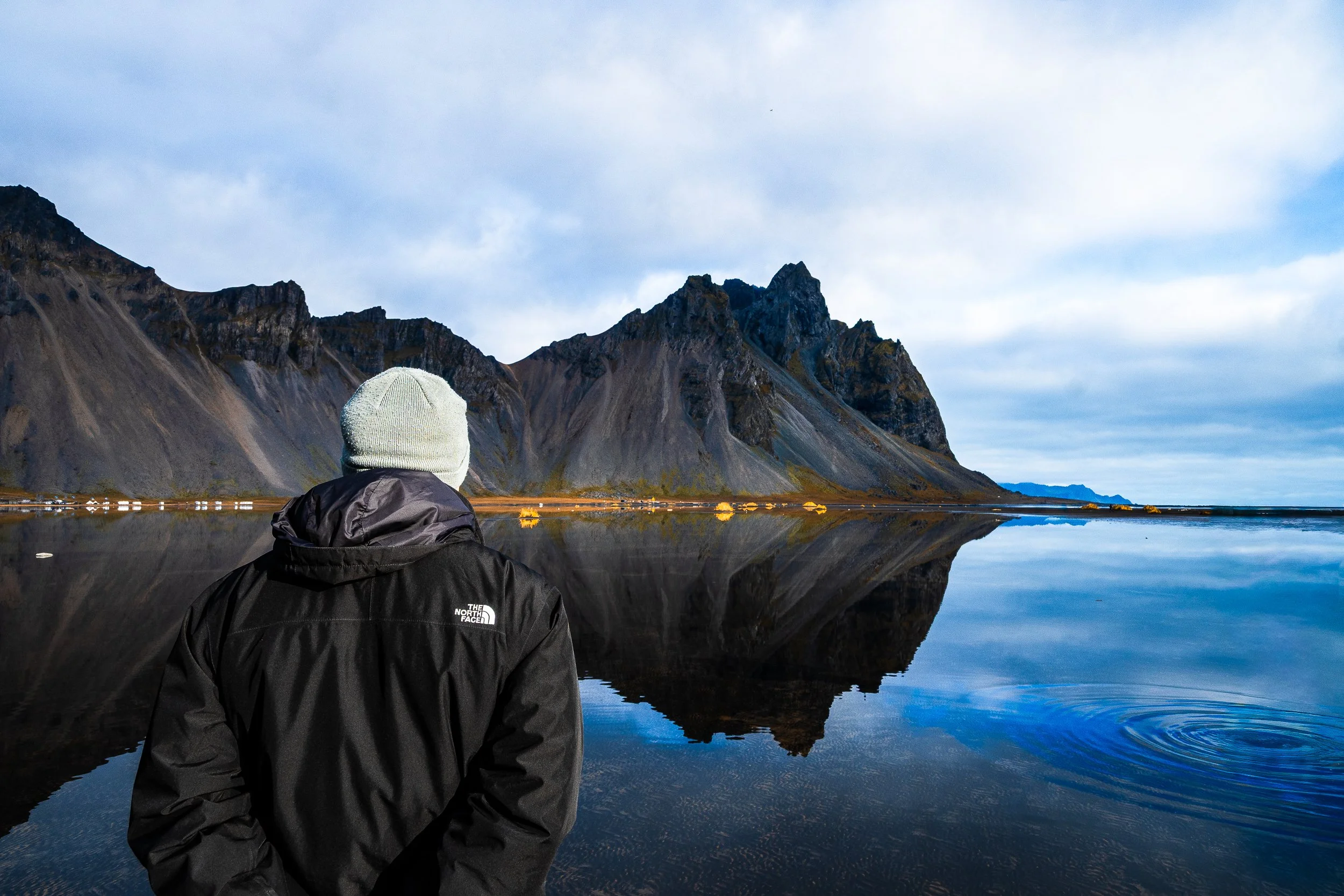 Person in a black jacket and knit hat viewing rugged mountains reflected in a calm Icelandic lake