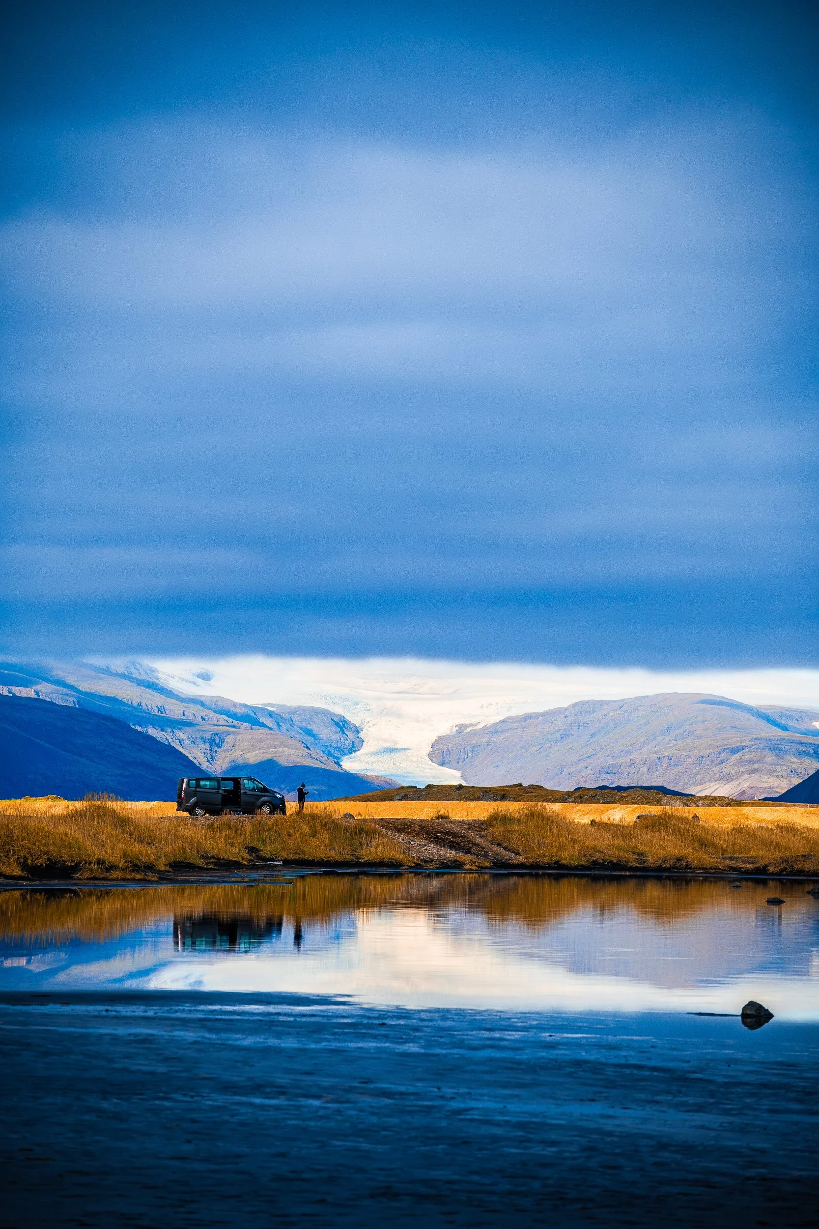Person standing beside a van overlooking a glacier-covered mountain valley and calm reflective lake